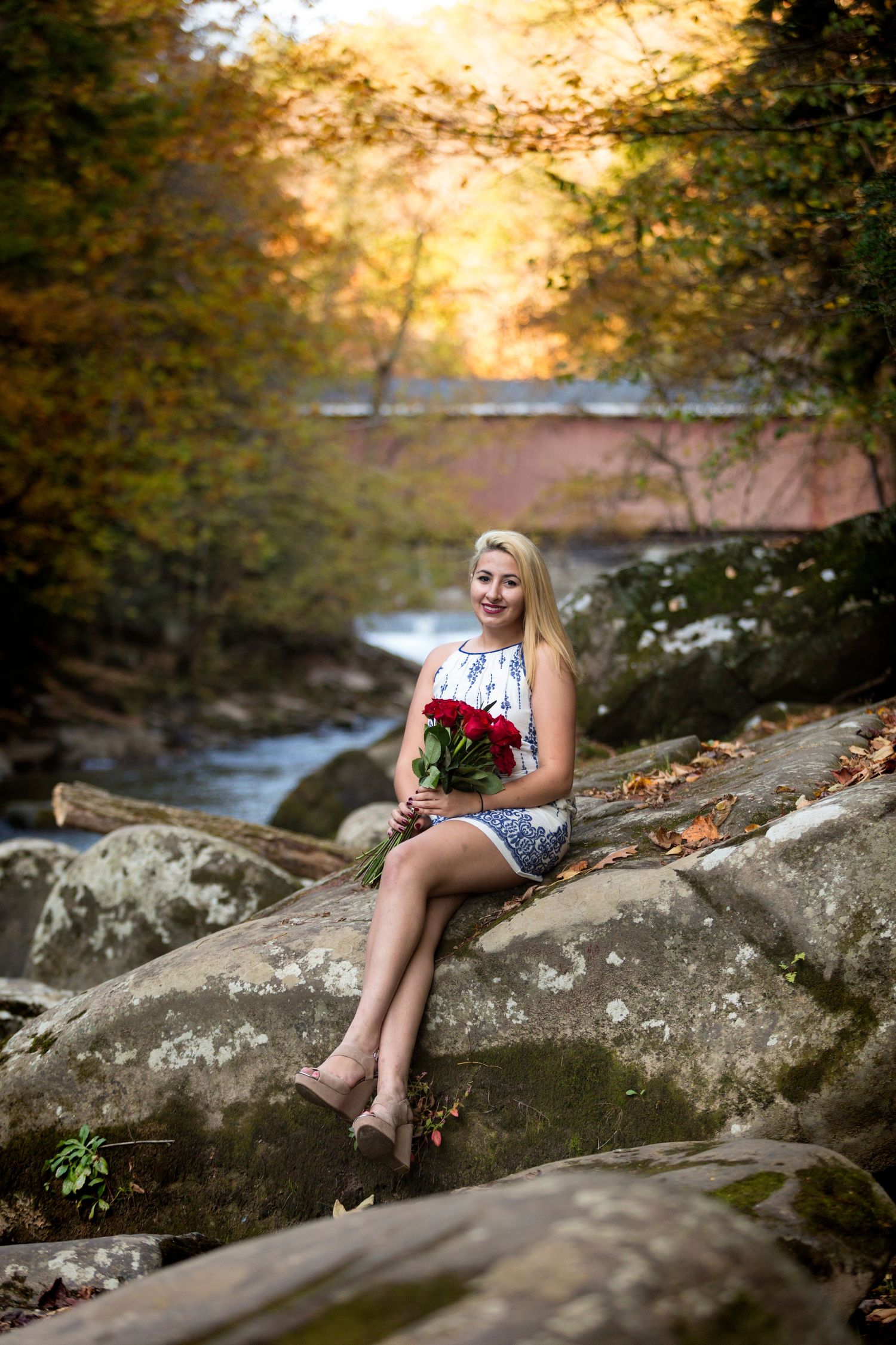 Smiling blonde woman sitting on rocks holding red roses bouquet near a stream with autumn foliage and covered bridge.