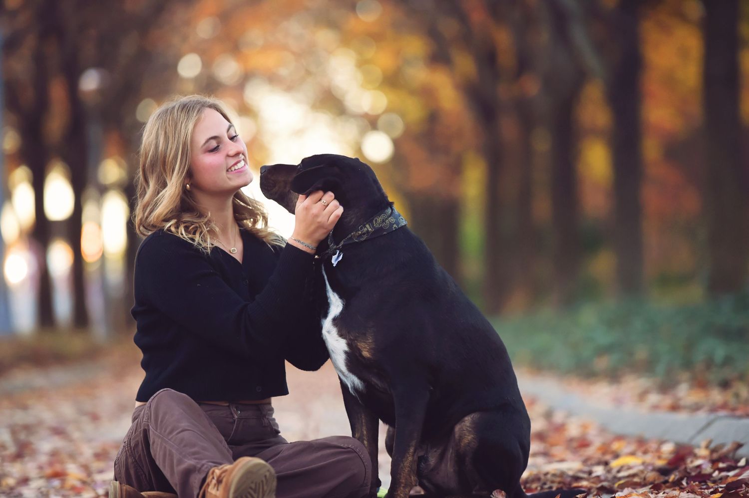 Smiling blonde woman sitting outdoors in autumn park, affectionately interacting with a black dog amid colorful fallen leaves.