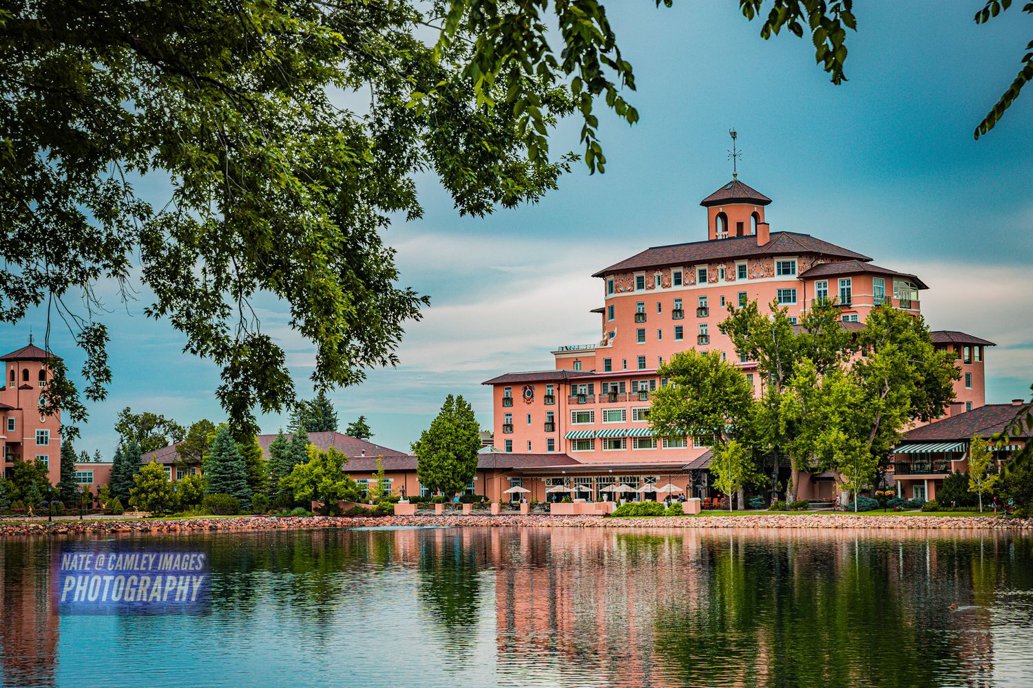 A panoramic view of Broadmoor Hotel's pink facade reflecting in Cheyenne Lake with lush trees framing the scene.