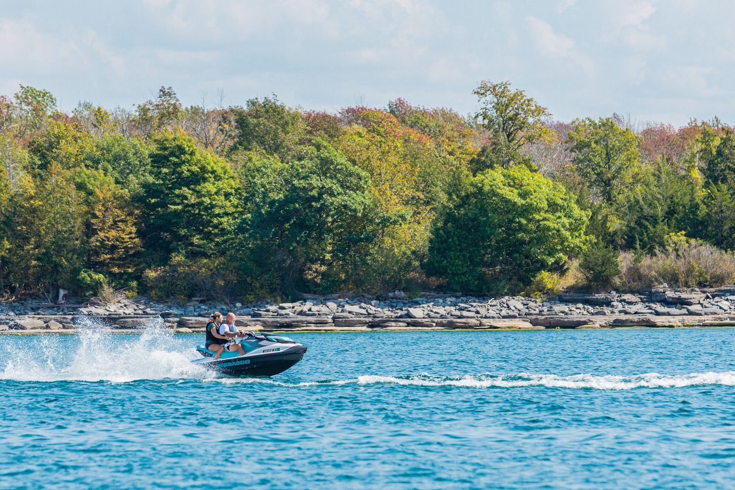 A high-speed boat races across blue waters creating dramatic wake spray near a rocky tree-lined shoreline.
