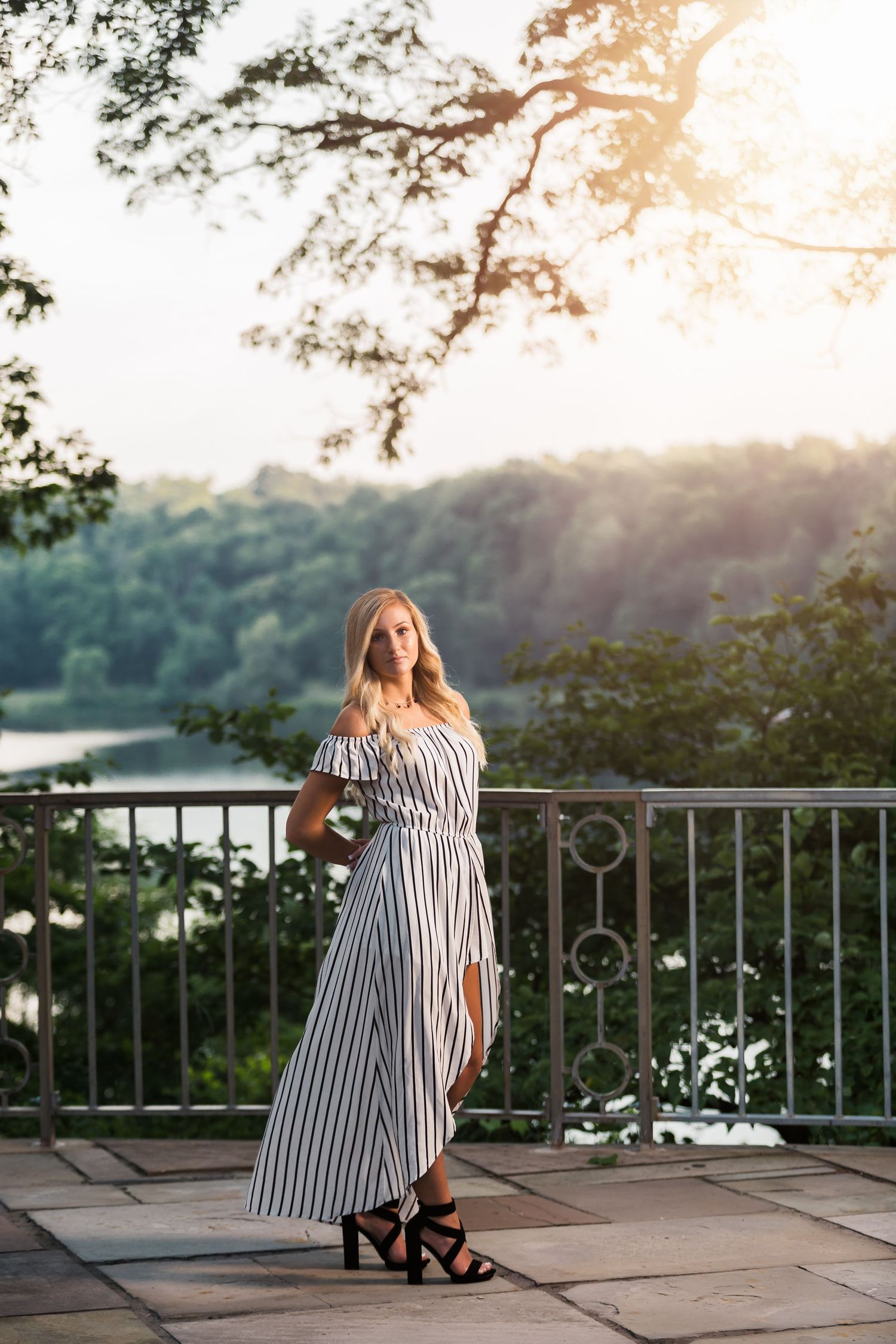 Blonde woman in striped off-shoulder maxi dress poses on stone terrace with lush green trees and river backdrop.