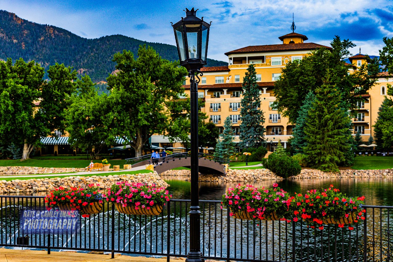 Luxury lakeside resort with ornate black gates and colorful flower gardens surrounded by mountain scenery.