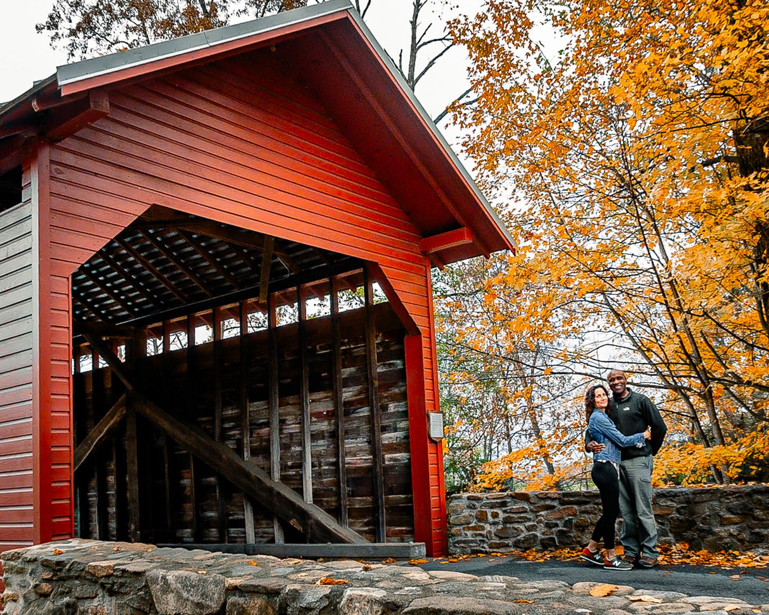 A red covered bridge walkway with stone walls and autumn foliage in the background.