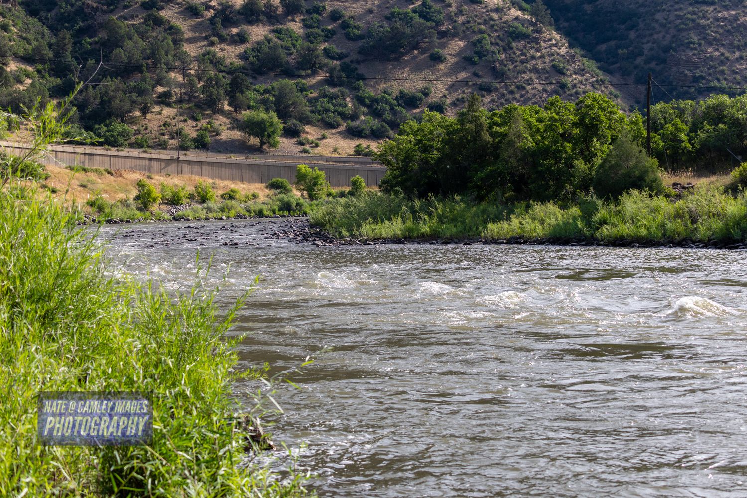 A scenic panoramic view of a rushing river surrounded by rocky terrain and green vegetation in a mountainous landscape.