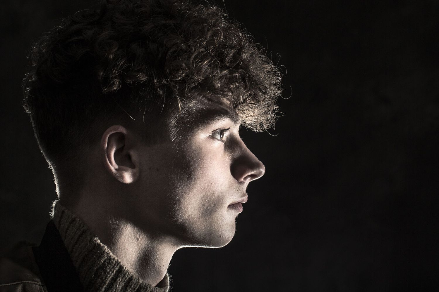 Side profile of a young man with curly hair dramatically lit against a dark background.