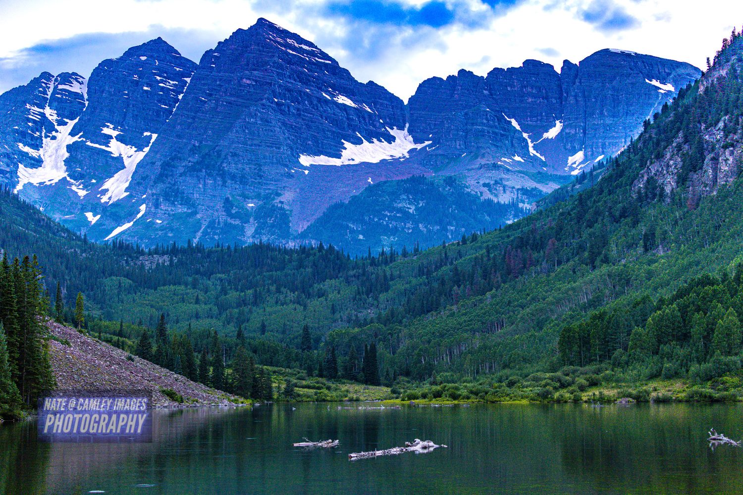 Snow-capped mountain peaks tower over a serene alpine lake surrounded by evergreen forest in Maroon Bells, Colorado.