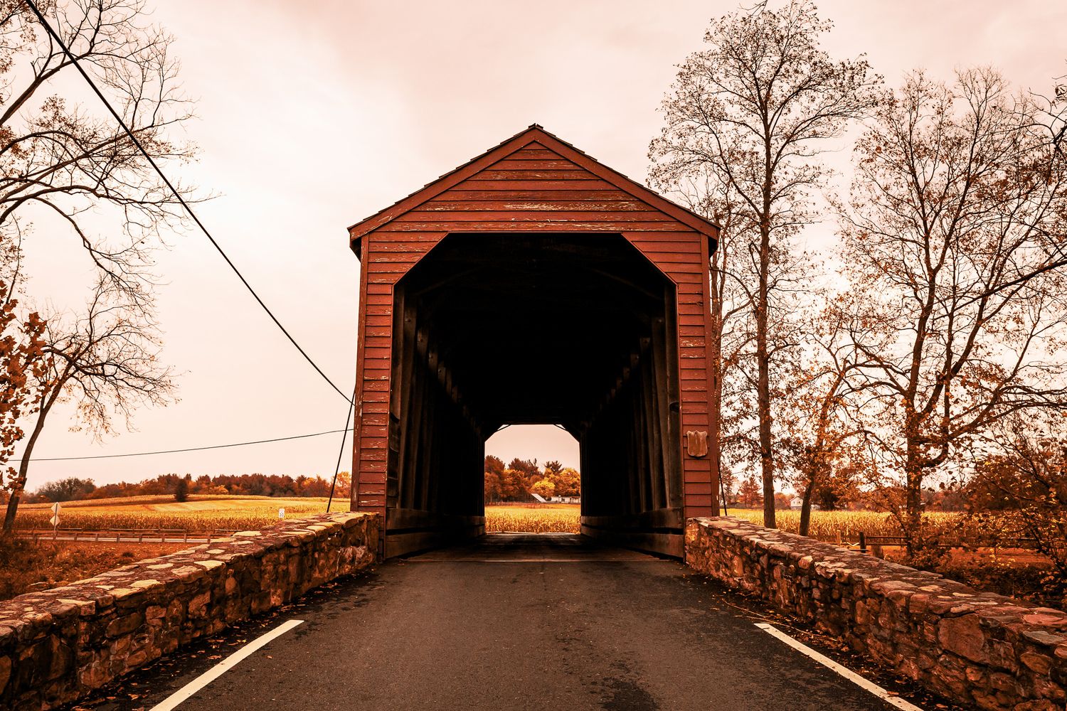 A rustic red covered bridge with stone walls stands against an autumn landscape at sunset.