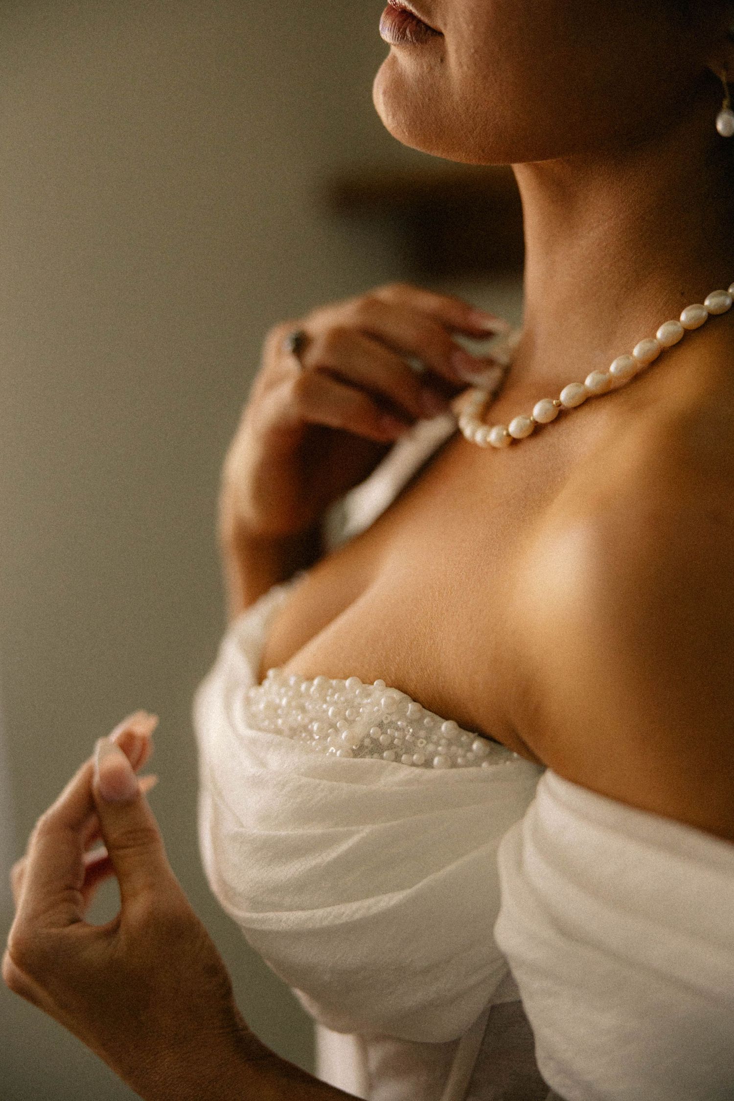 Bride in white beaded strapless gown adjusting pearl necklace, elegant bridal jewelry and dress detail.