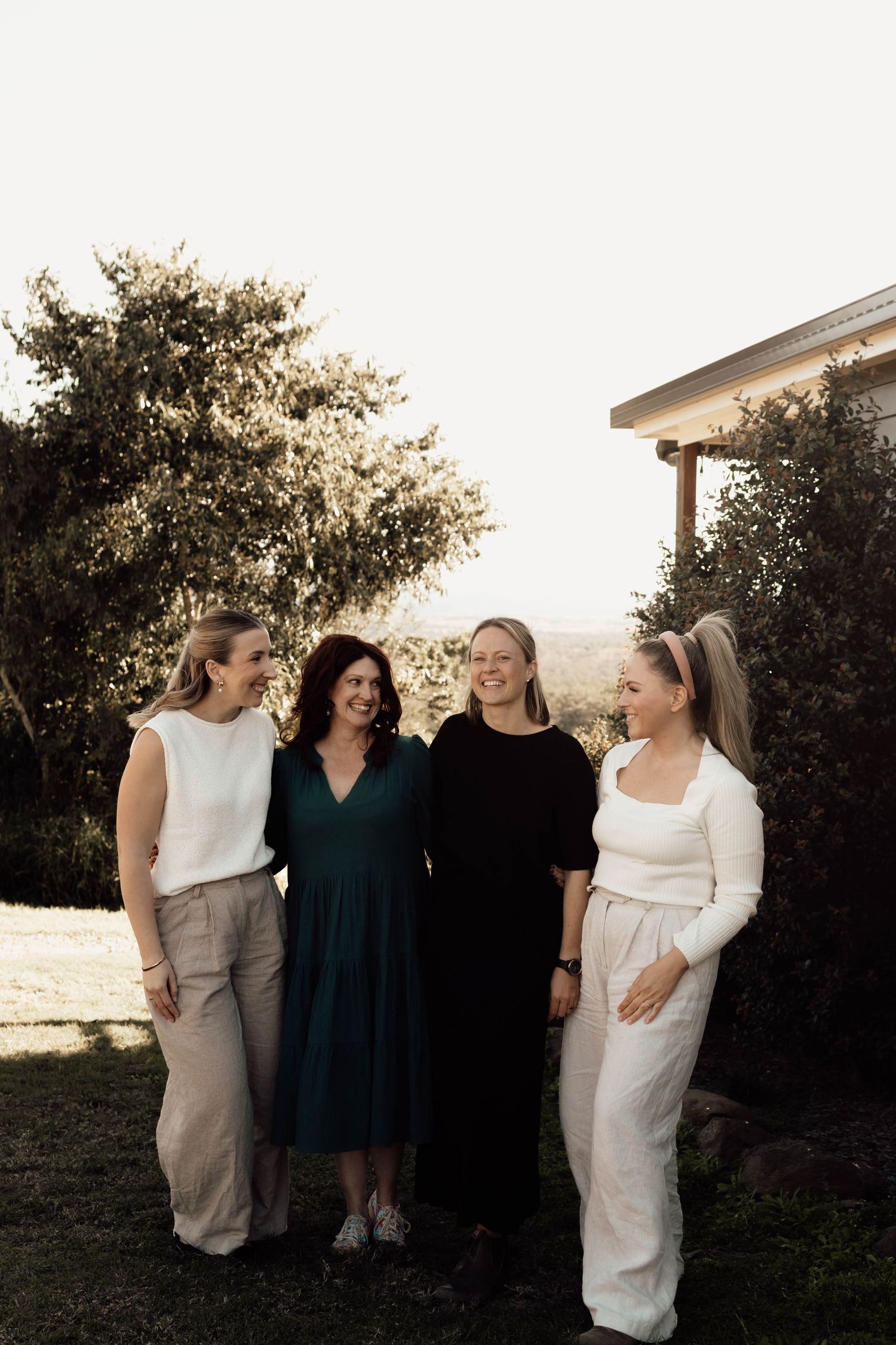 Four women smiling together outdoors in a garden setting, wearing elegant neutral and dark toned outfits.