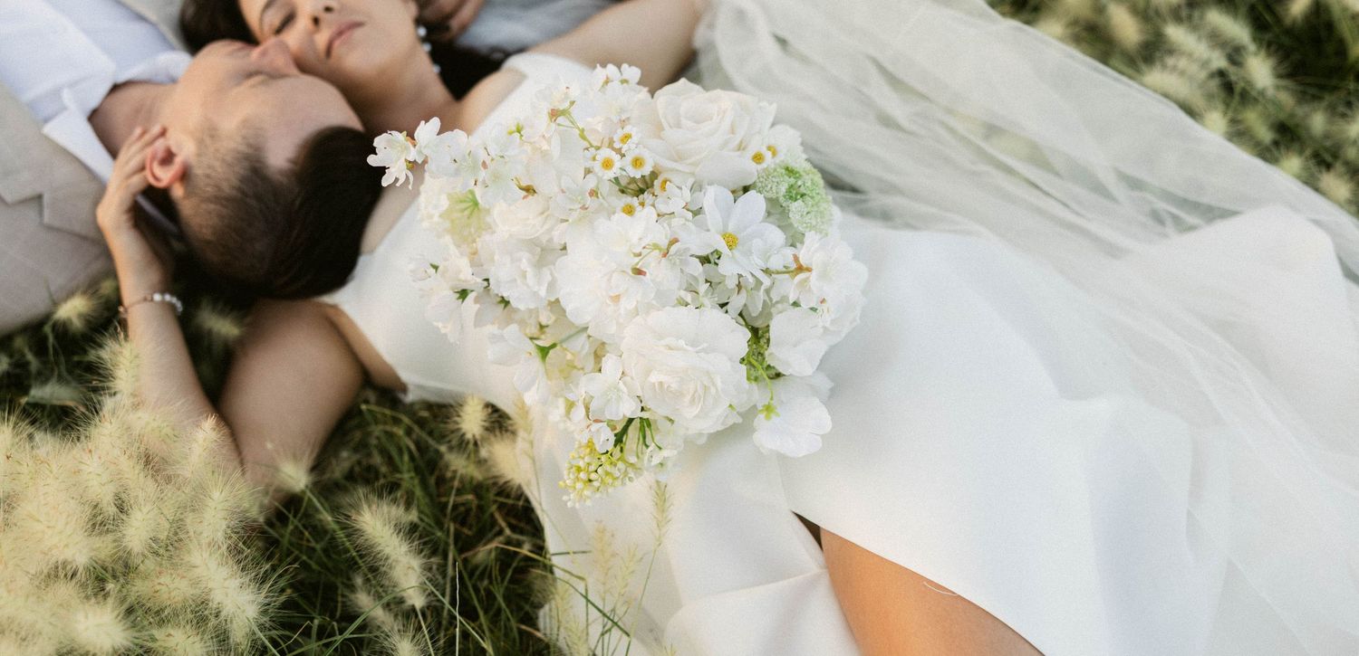 Bride and groom lying in grass, bride holding white floral bouquet with roses in elegant wedding dress.