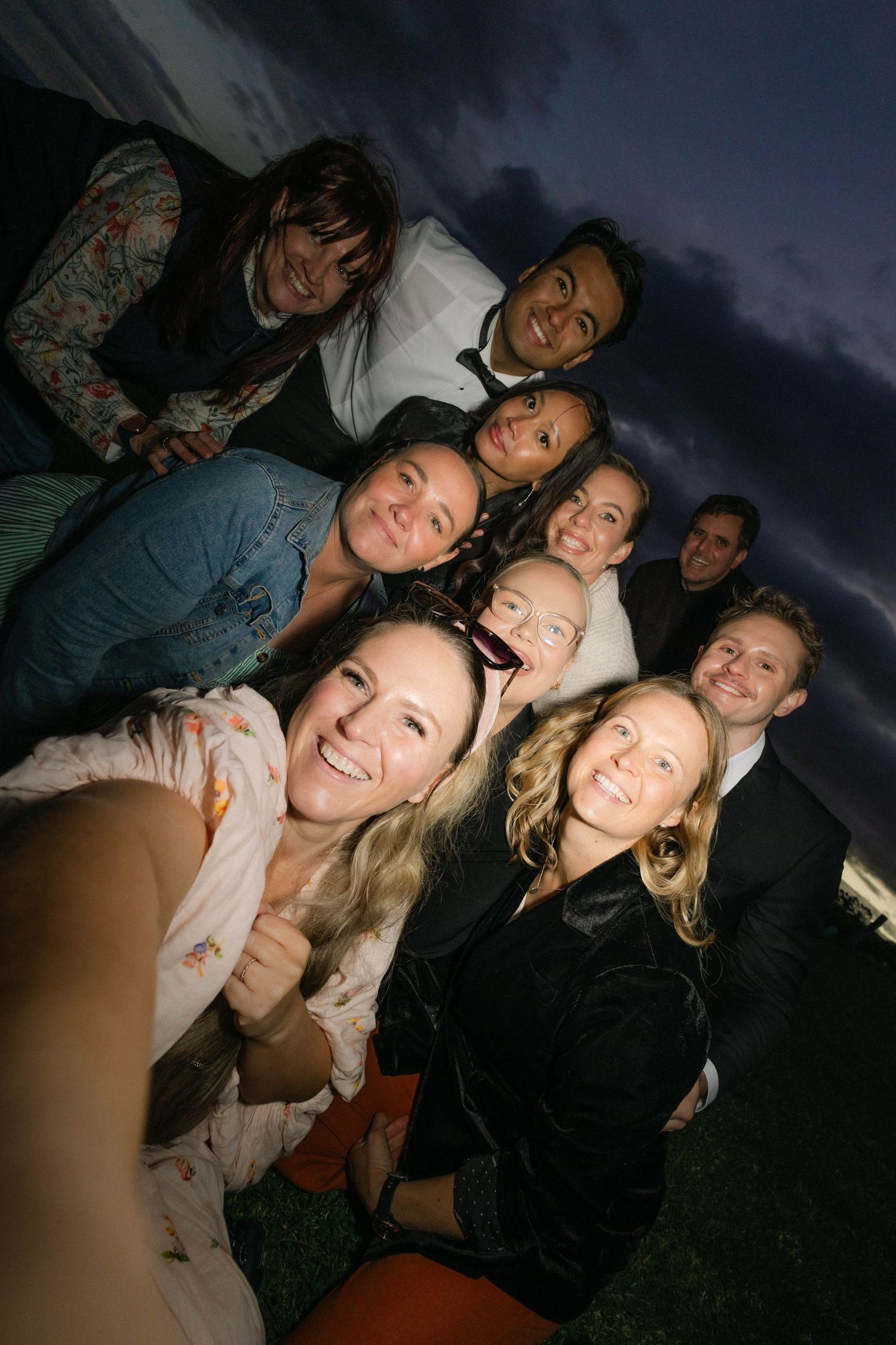 Group of smiling friends taking a fun outdoor selfie together at dusk under a dramatic cloudy sky.