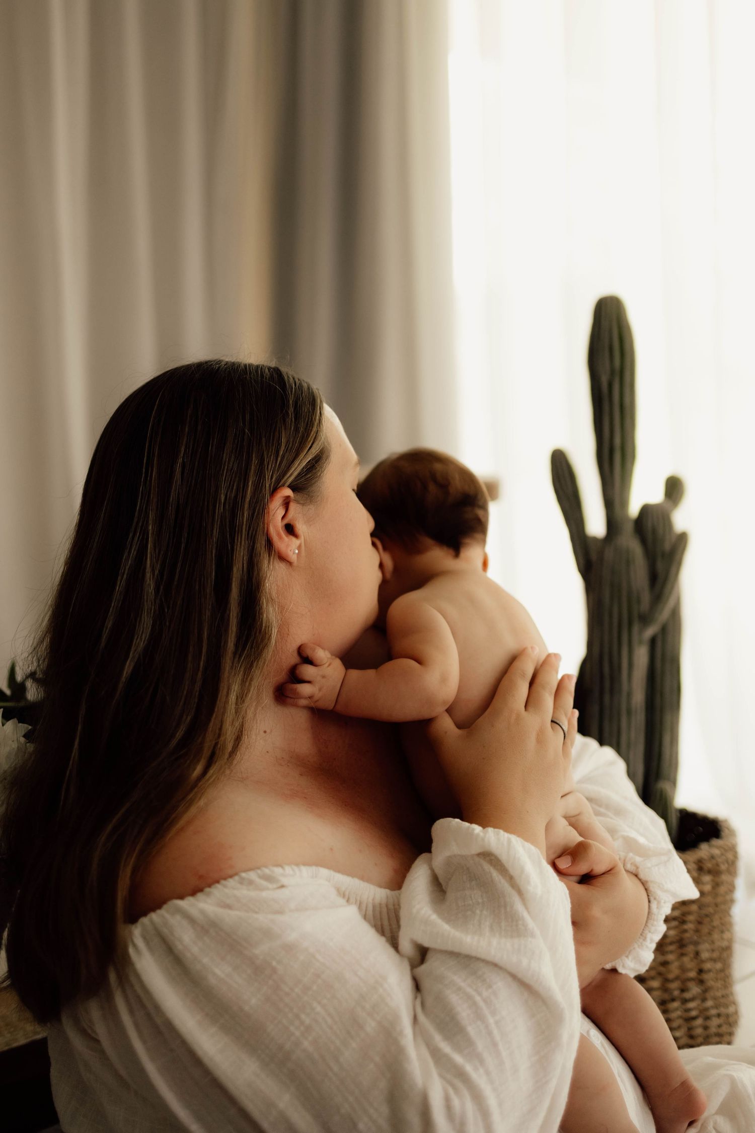 A mother in a white off-shoulder top tenderly kisses her baby while holding them near a cactus by a bright window.