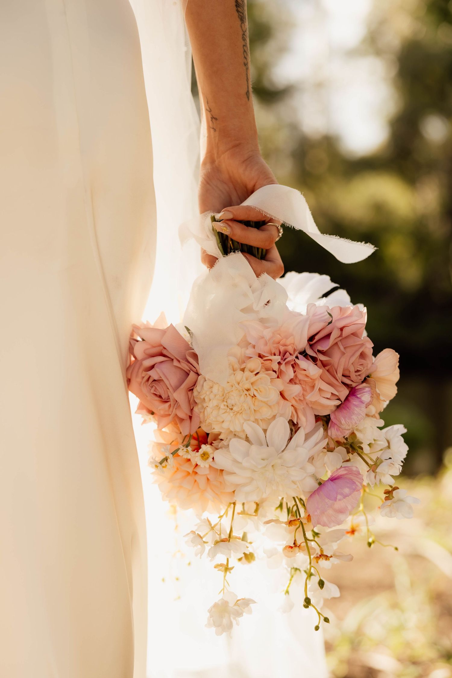 Bride holding a romantic bridal bouquet of blush pink roses, white dahlias, and peach flowers with white ribbon.