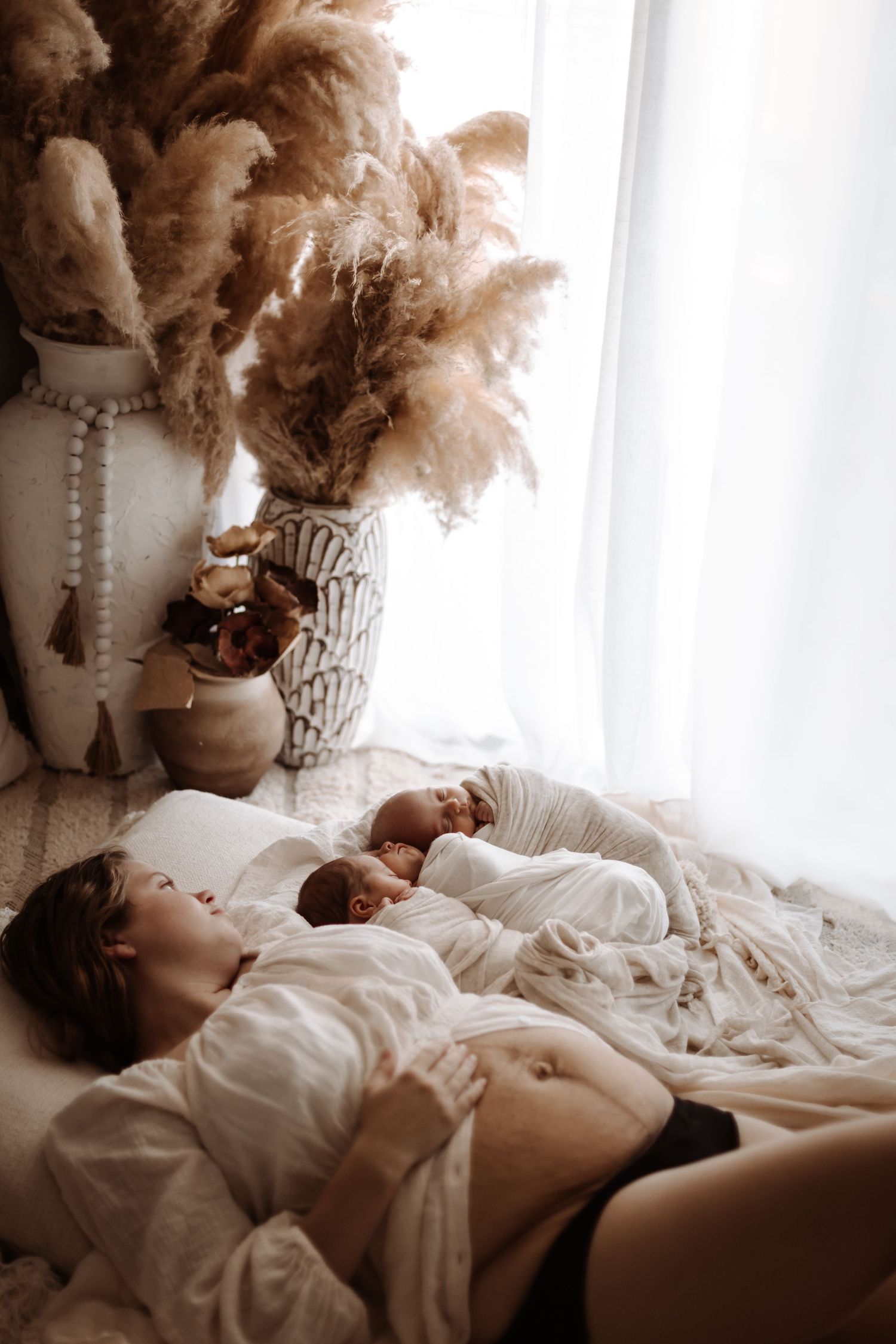 Mother resting on bed with newborn twins wrapped in white blankets near boho vase with pampas grass.