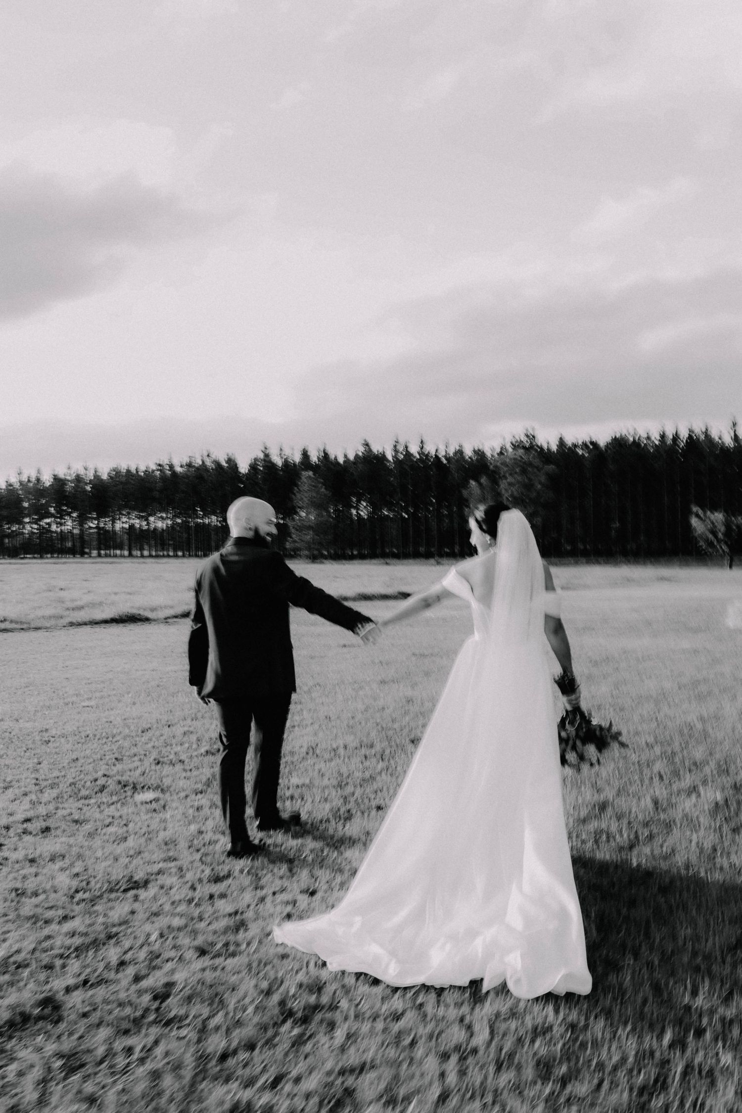 Black and white photo of a bride and groom holding hands in a field with trees in the background.