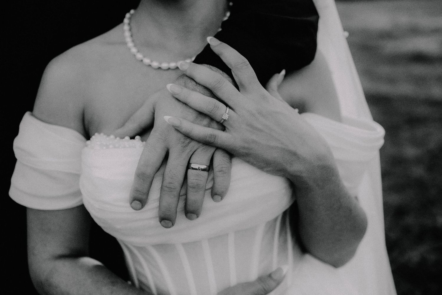 Black and white wedding photo of bride and groom embracing, showcasing diamond rings and pearl necklace.