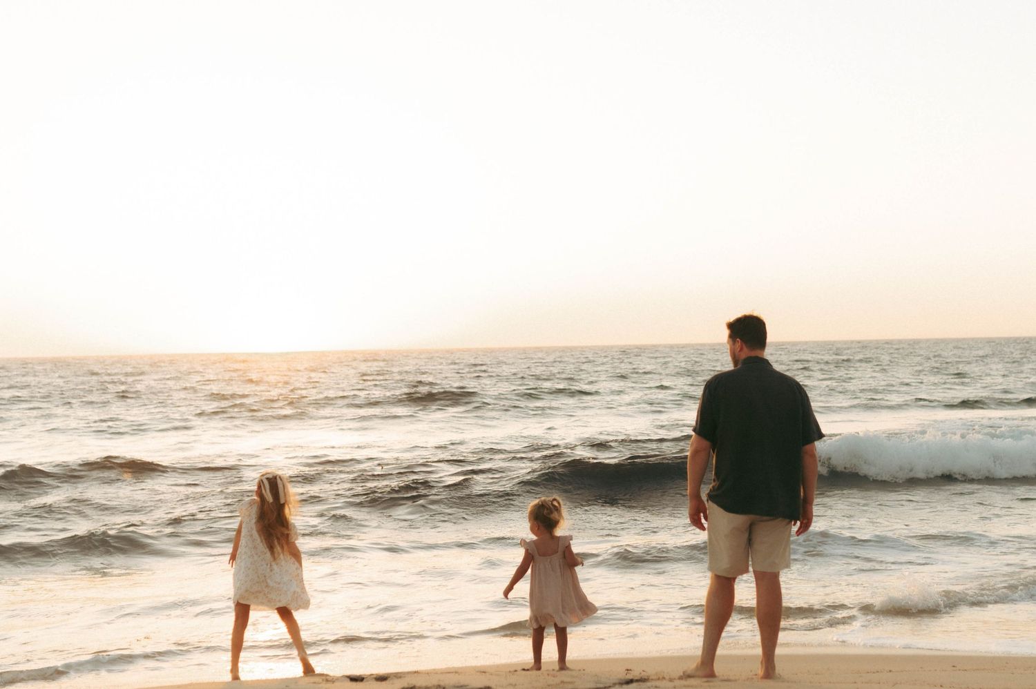 Father and two young daughters standing on a sandy beach watching ocean waves at sunset.