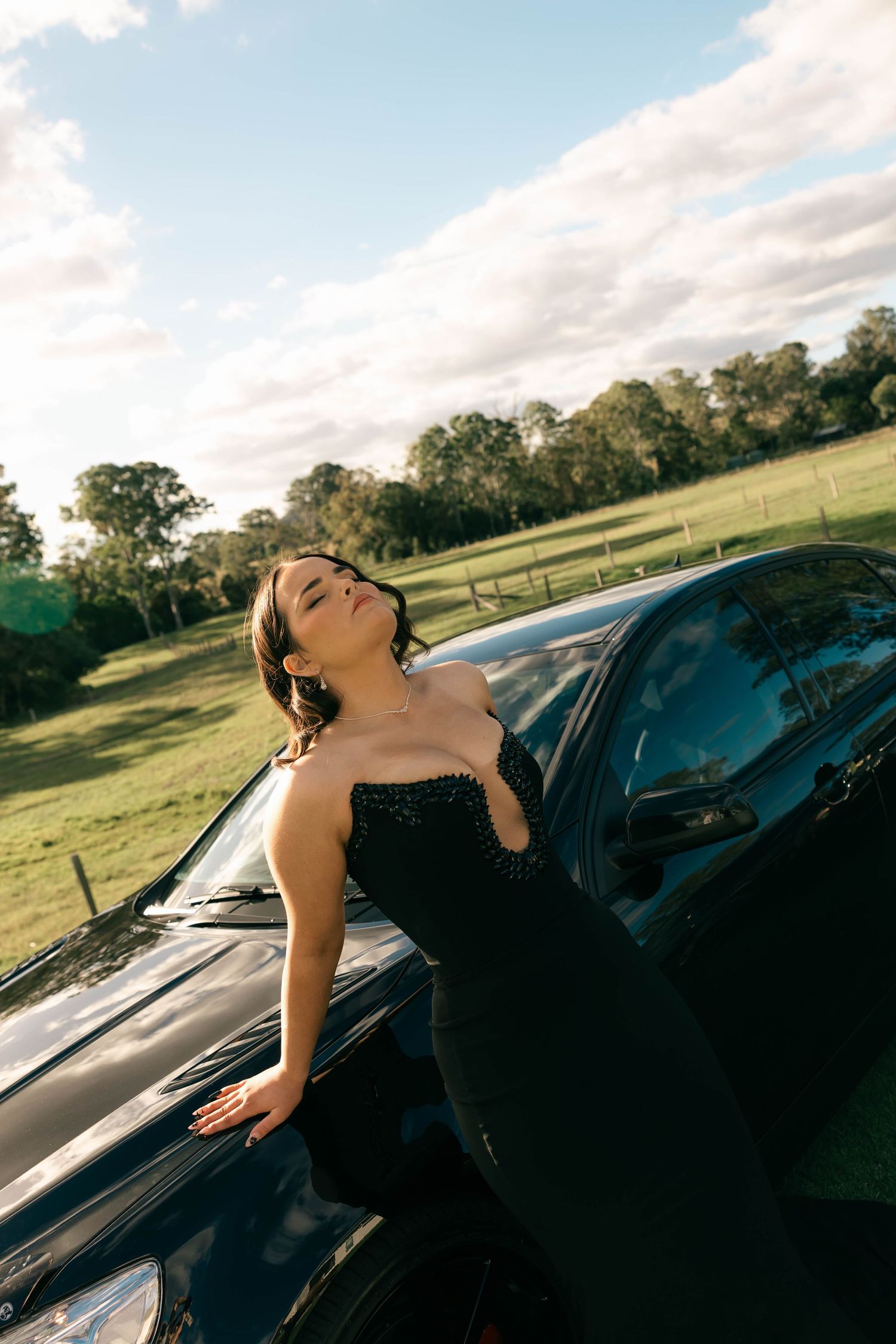 Woman in black evening gown leaning against dark car in scenic green field under cloudy sky.