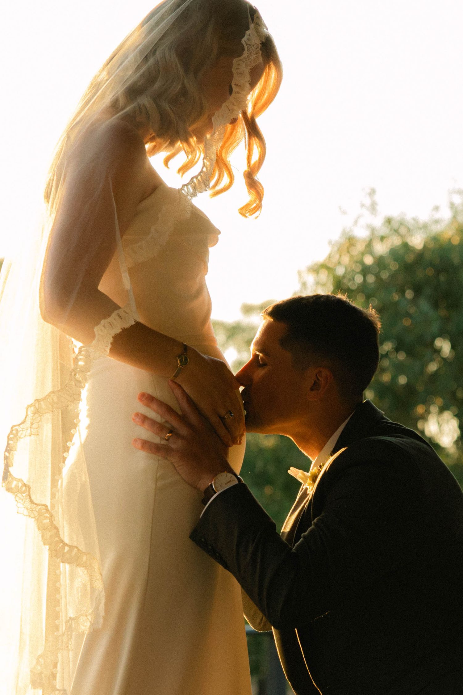 Groom kneeling to kiss bride's belly in golden sunset light, both in wedding attire with veil flowing.