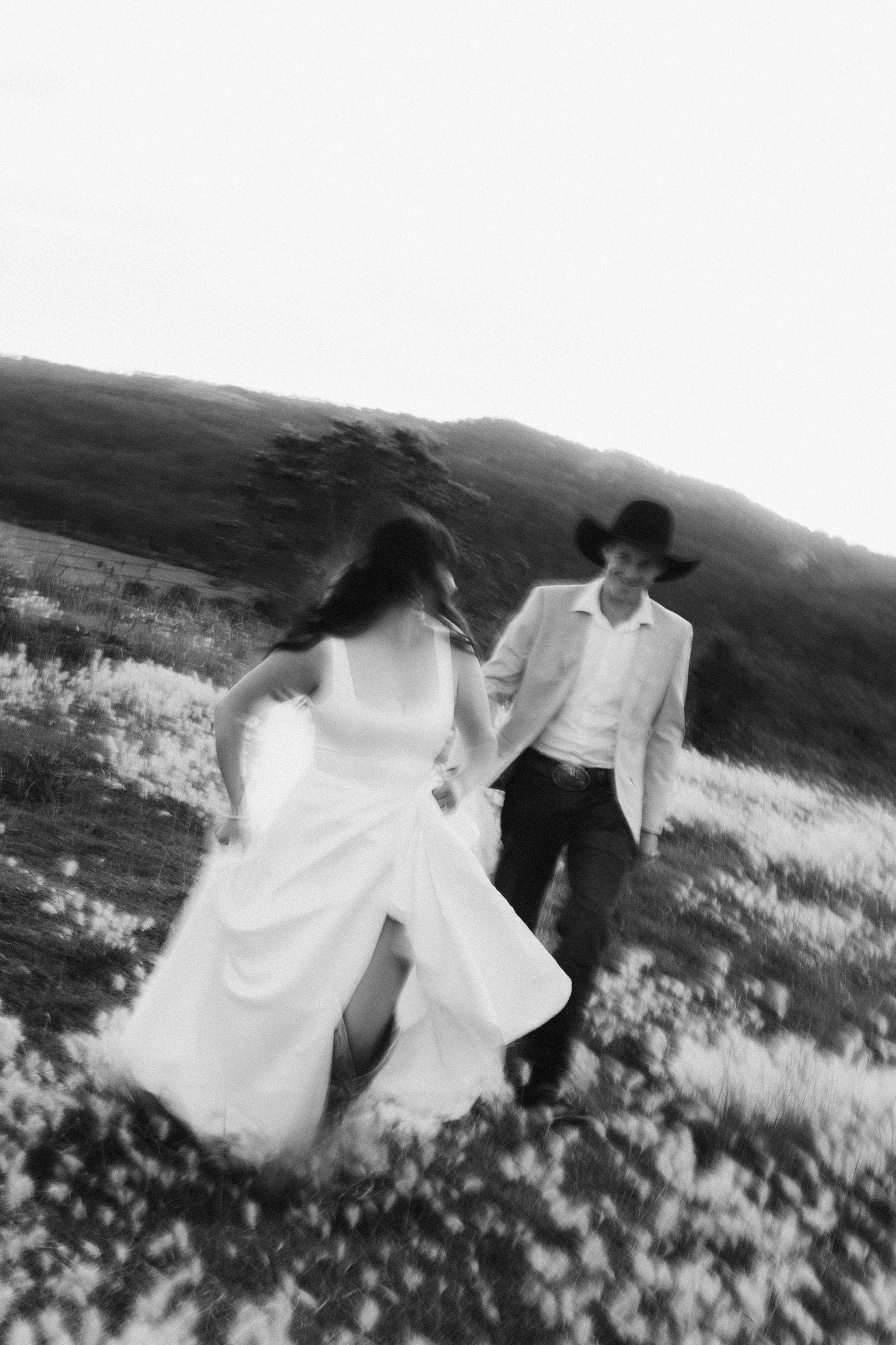 Black and white photo of a bride in white dress and groom in cowboy hat running through a flower field.