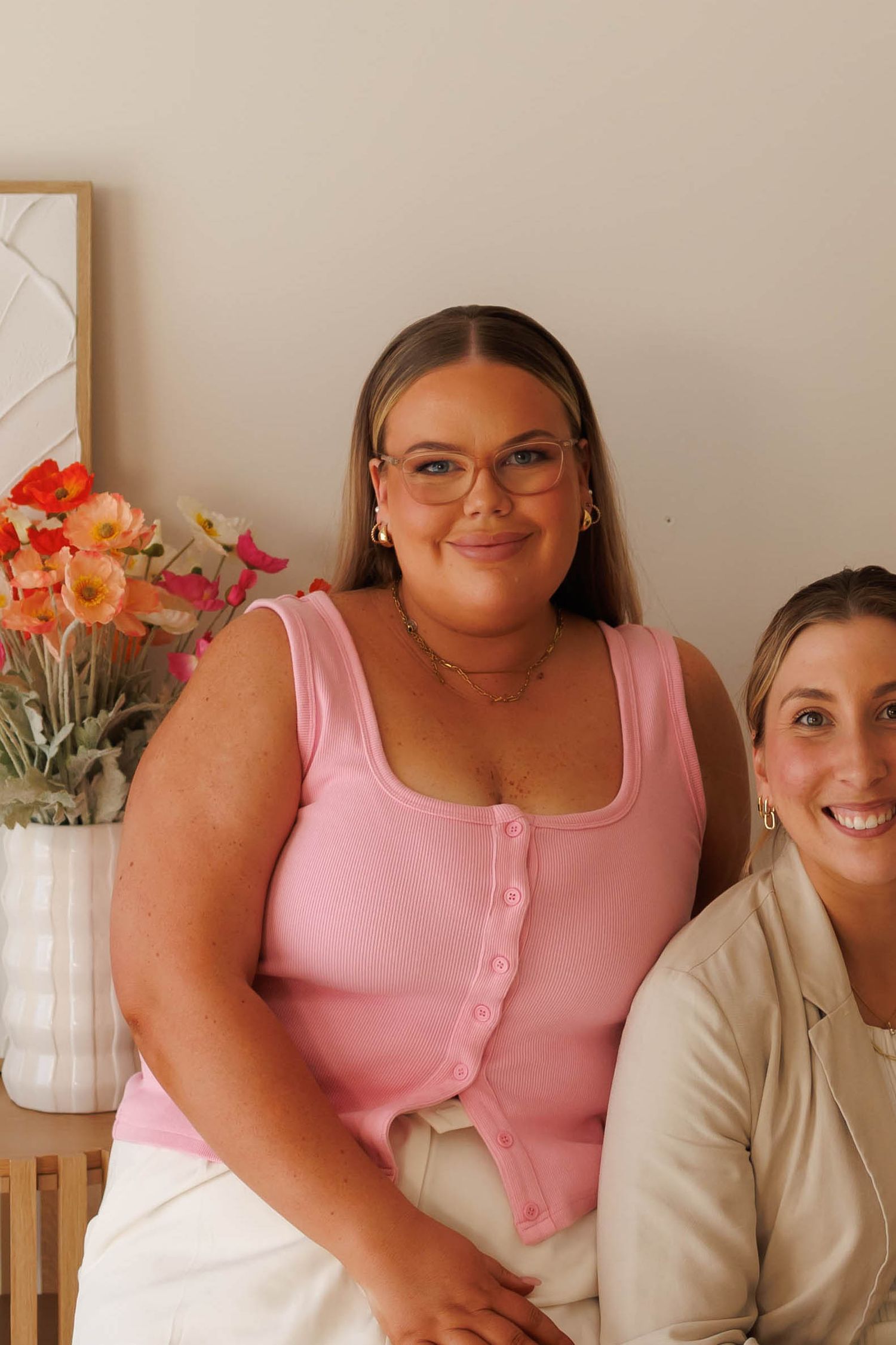 Two women sitting together, one wearing pink ribbed tank top with glasses, another in cream blazer, with colorful flowers behind.
