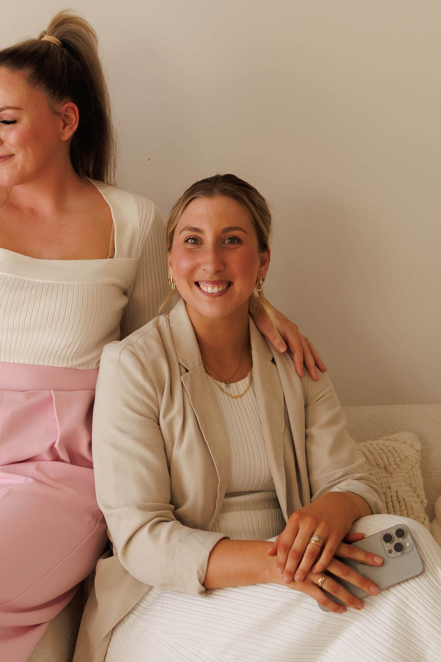 Two smiling women sitting together, one in a beige blazer holding a phone, the other in pink pants and white top.