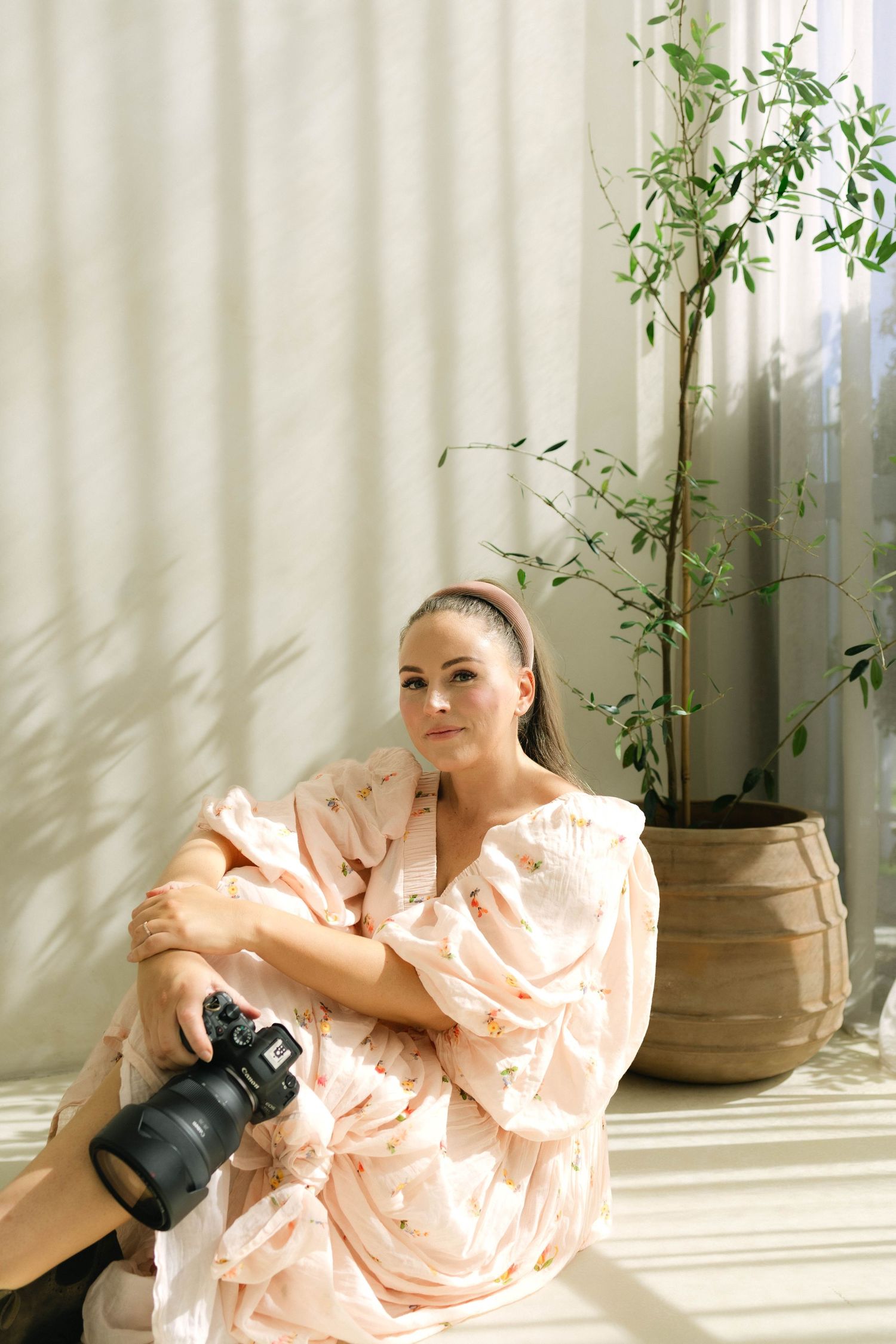 Woman in floral peach dress sitting on floor holding camera near potted plant by curtained window.
