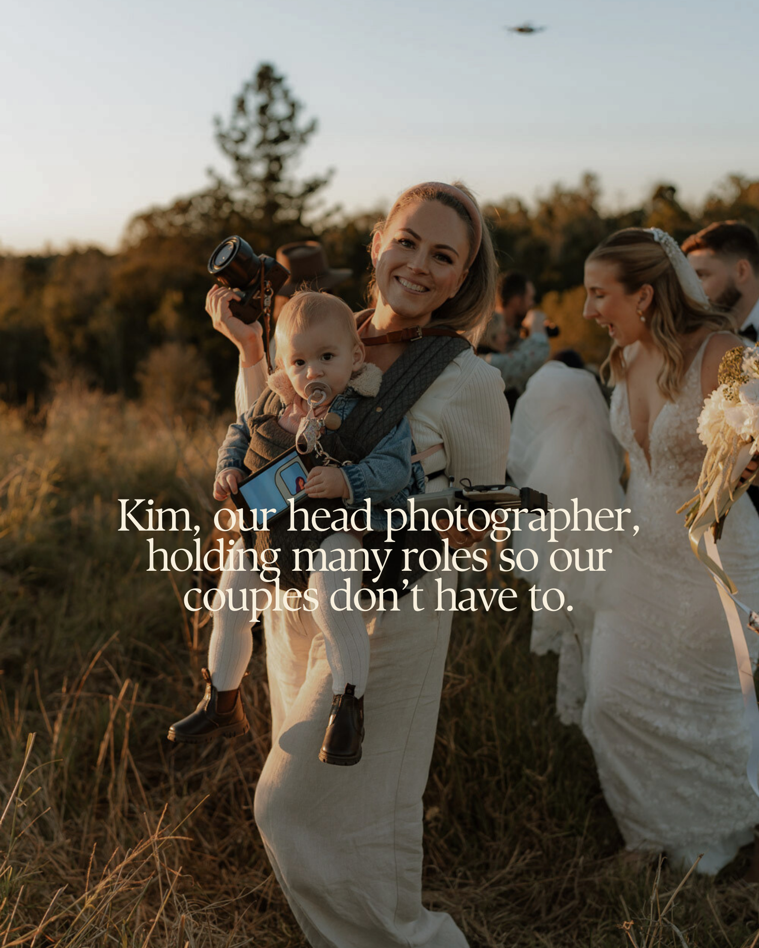 Kim, head wedding photographer, smiling while carrying a baby in a carrier and holding a camera at an outdoor wedding.