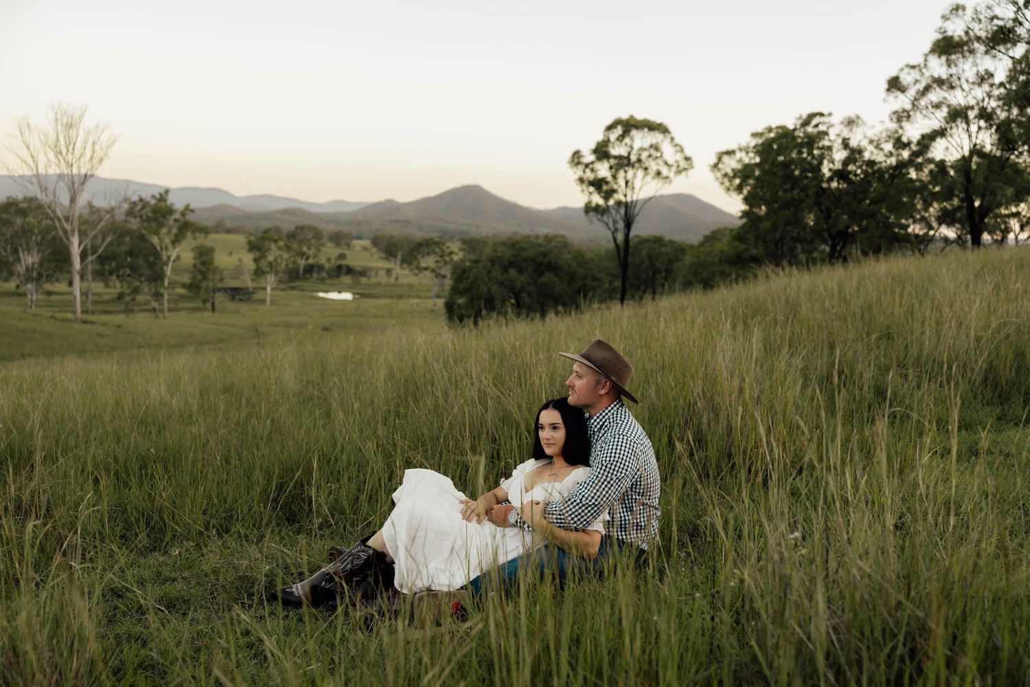 Couple sitting together in lush green field with mountain backdrop at dusk in rural Australia.