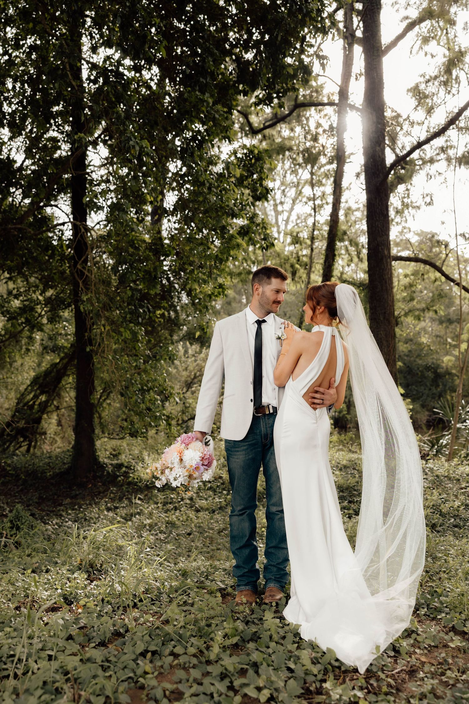 Bride and groom embrace in a lush forest setting, she in a backless white gown with veil, he in a white blazer and jeans.