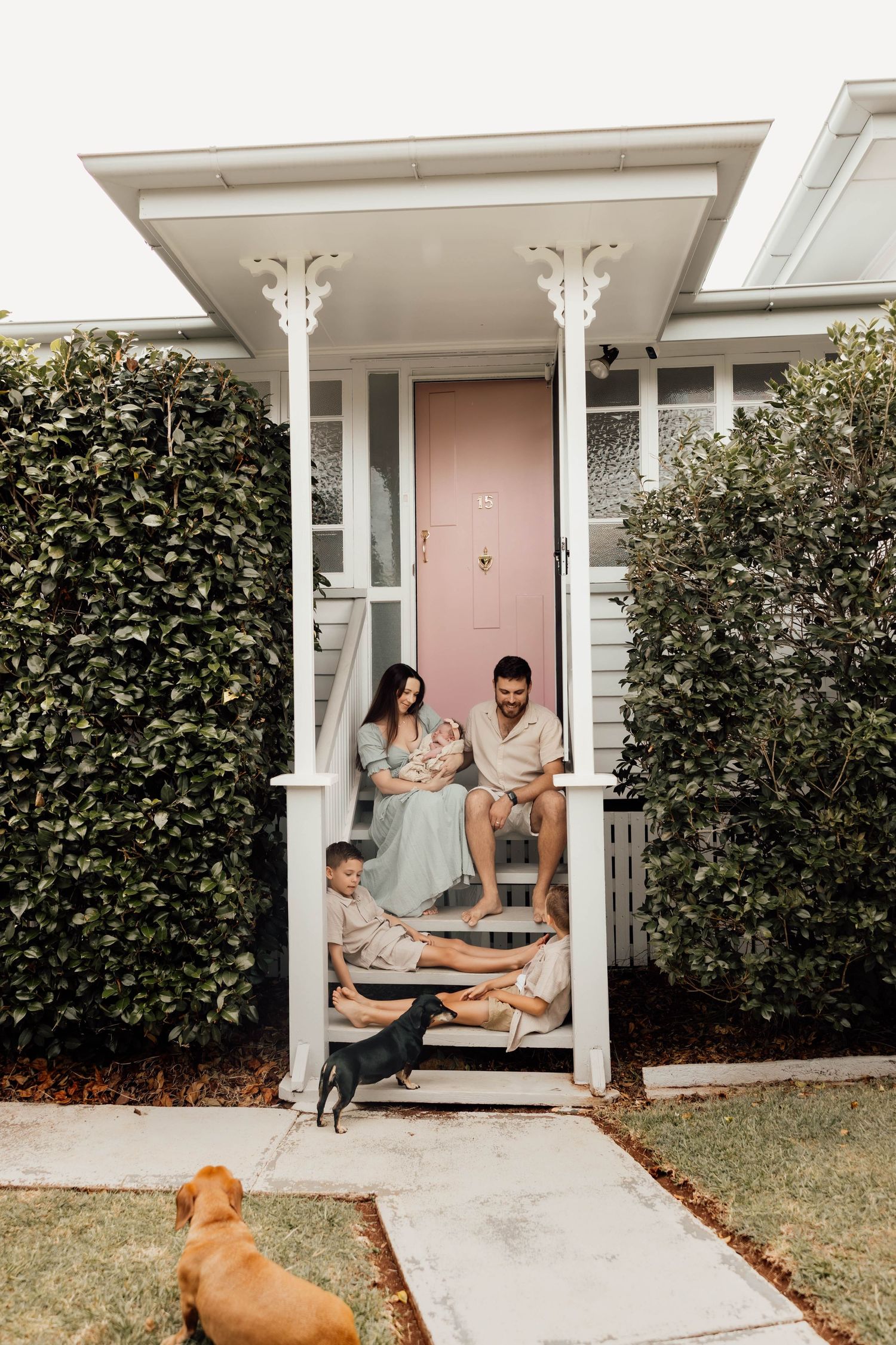 Family with newborn, toddler, and two dogs sitting on front porch steps of charming home with pink door.