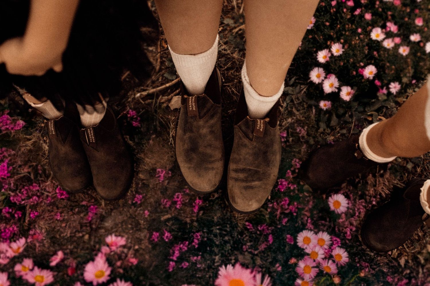 Overhead view of three people wearing brown boots standing among pink and purple wildflowers on dark soil.