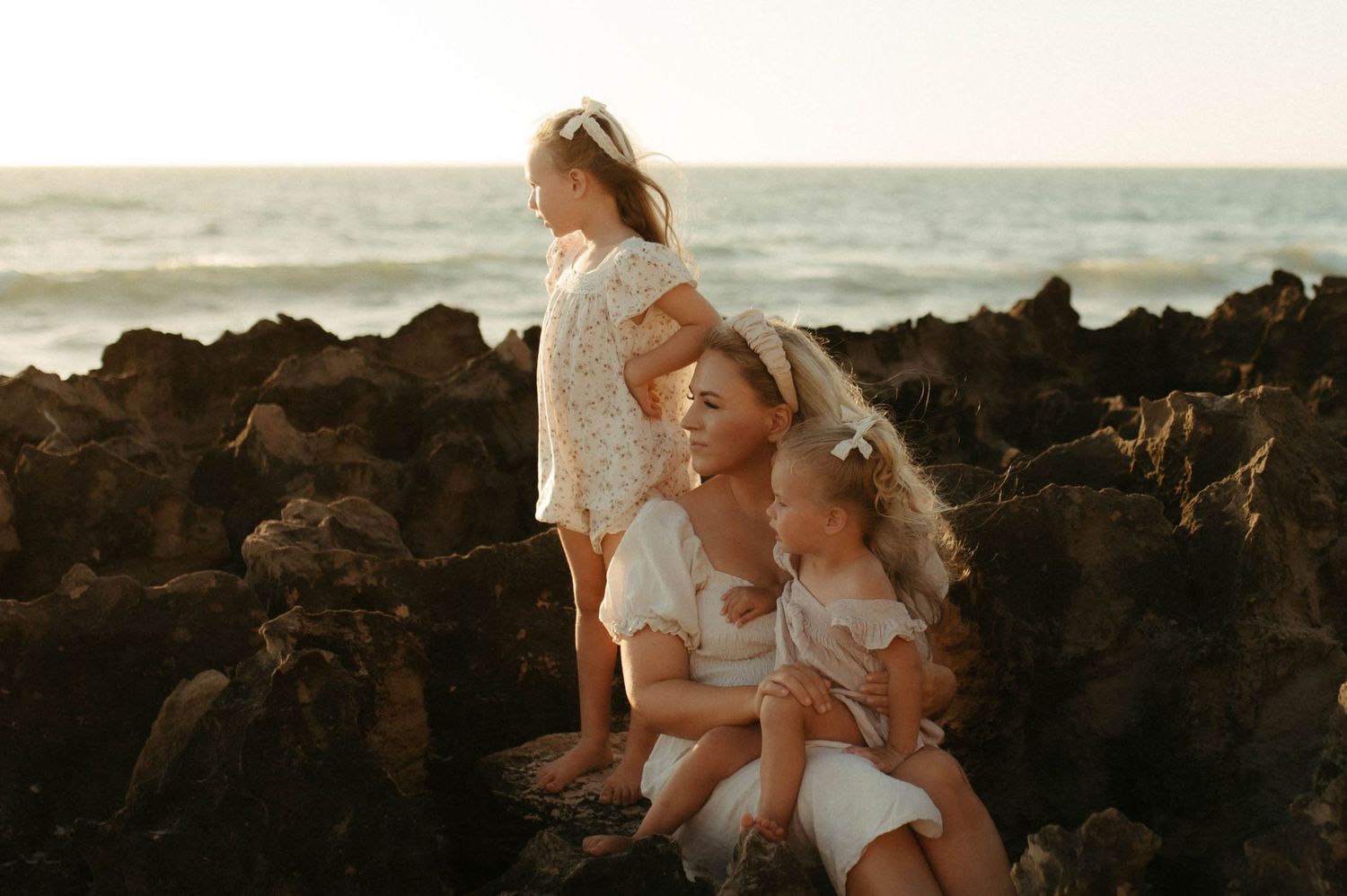 A mother and two young daughters in white dresses sit on rocky shore at golden hour with ocean waves behind them.
