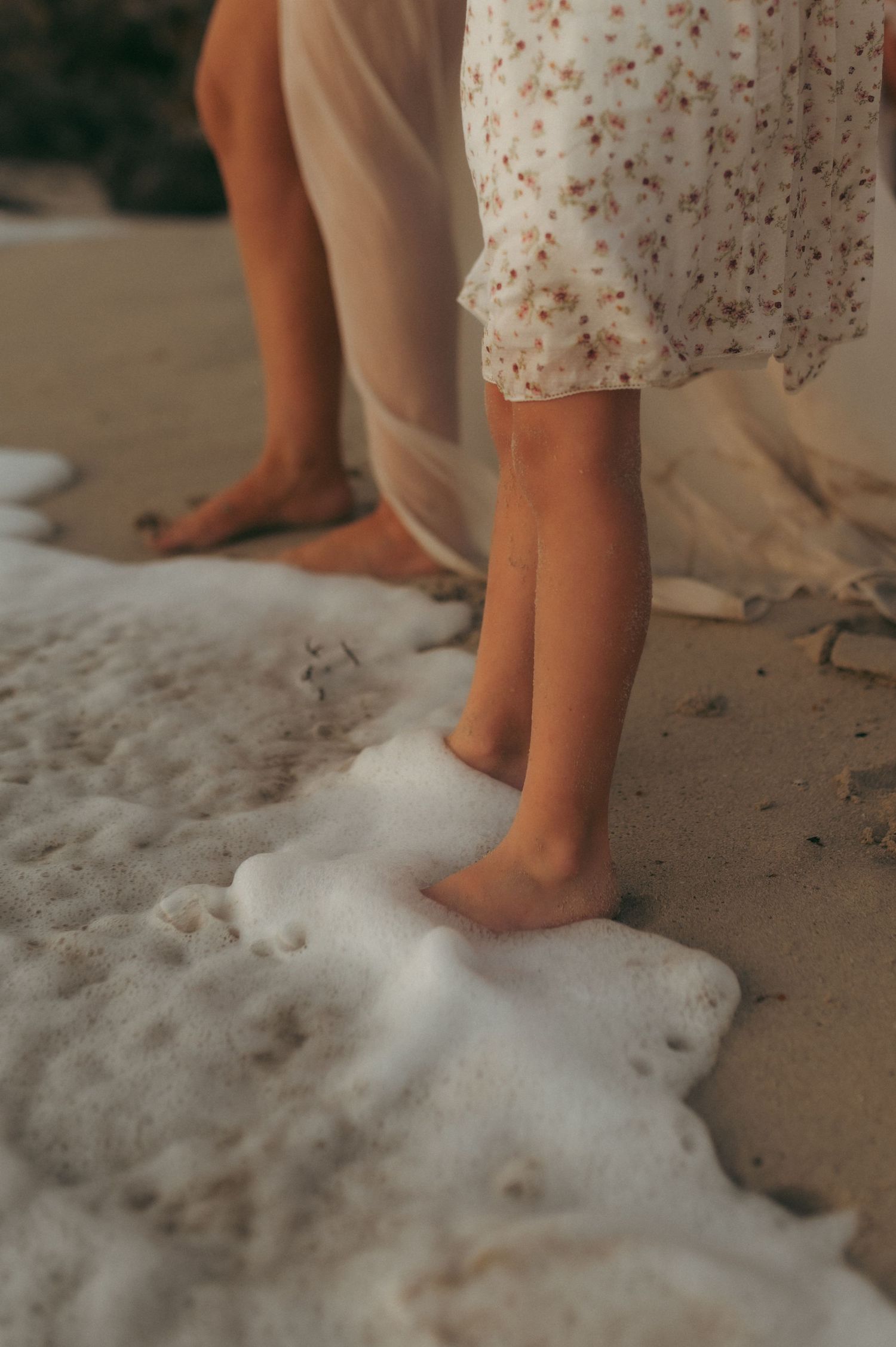 Two people standing barefoot on a sandy beach as gentle ocean waves wash over their feet.