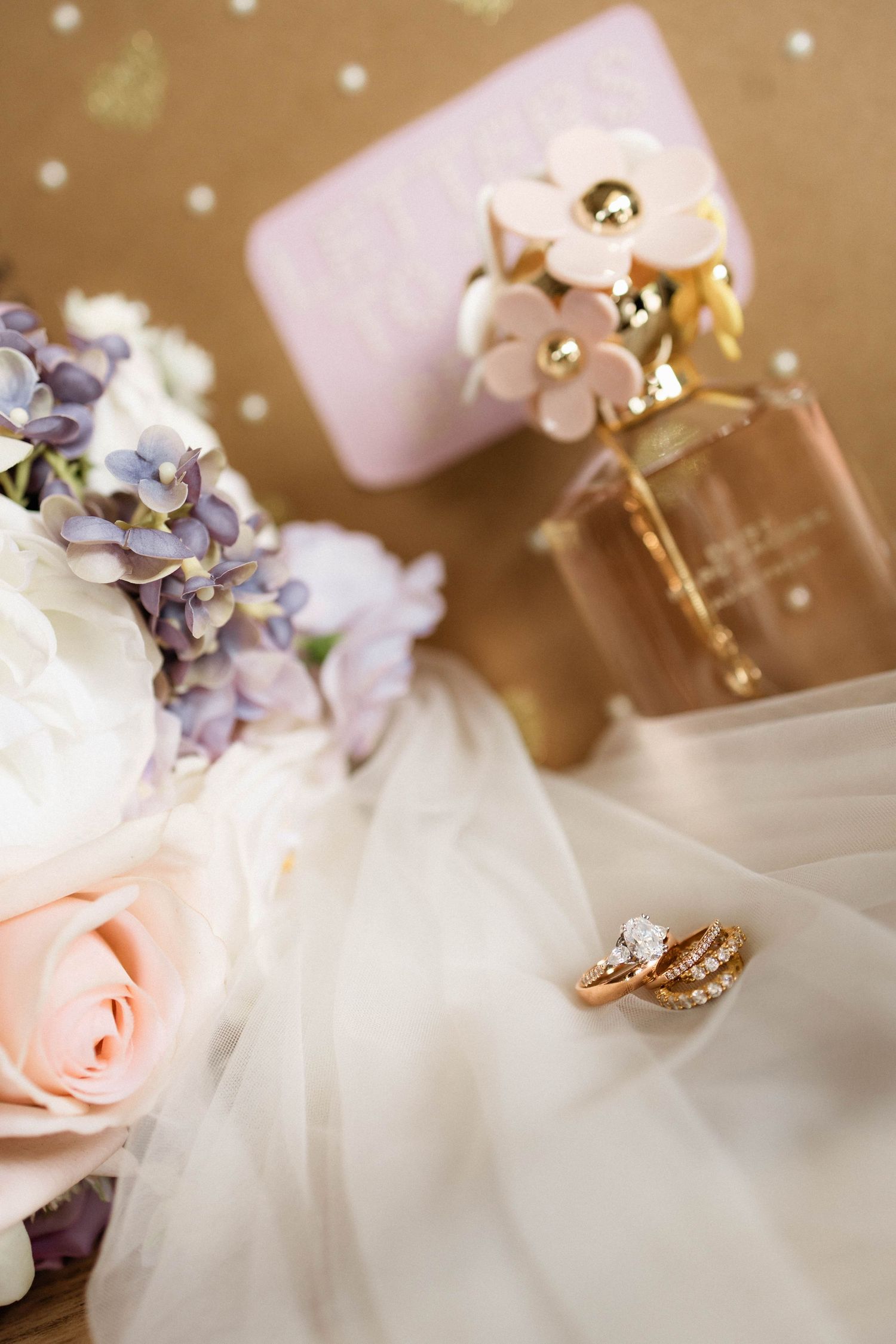 Wedding flat lay with gold rings, bridal bouquet of pink and white roses, and Marc Jacobs perfume on ivory veil.