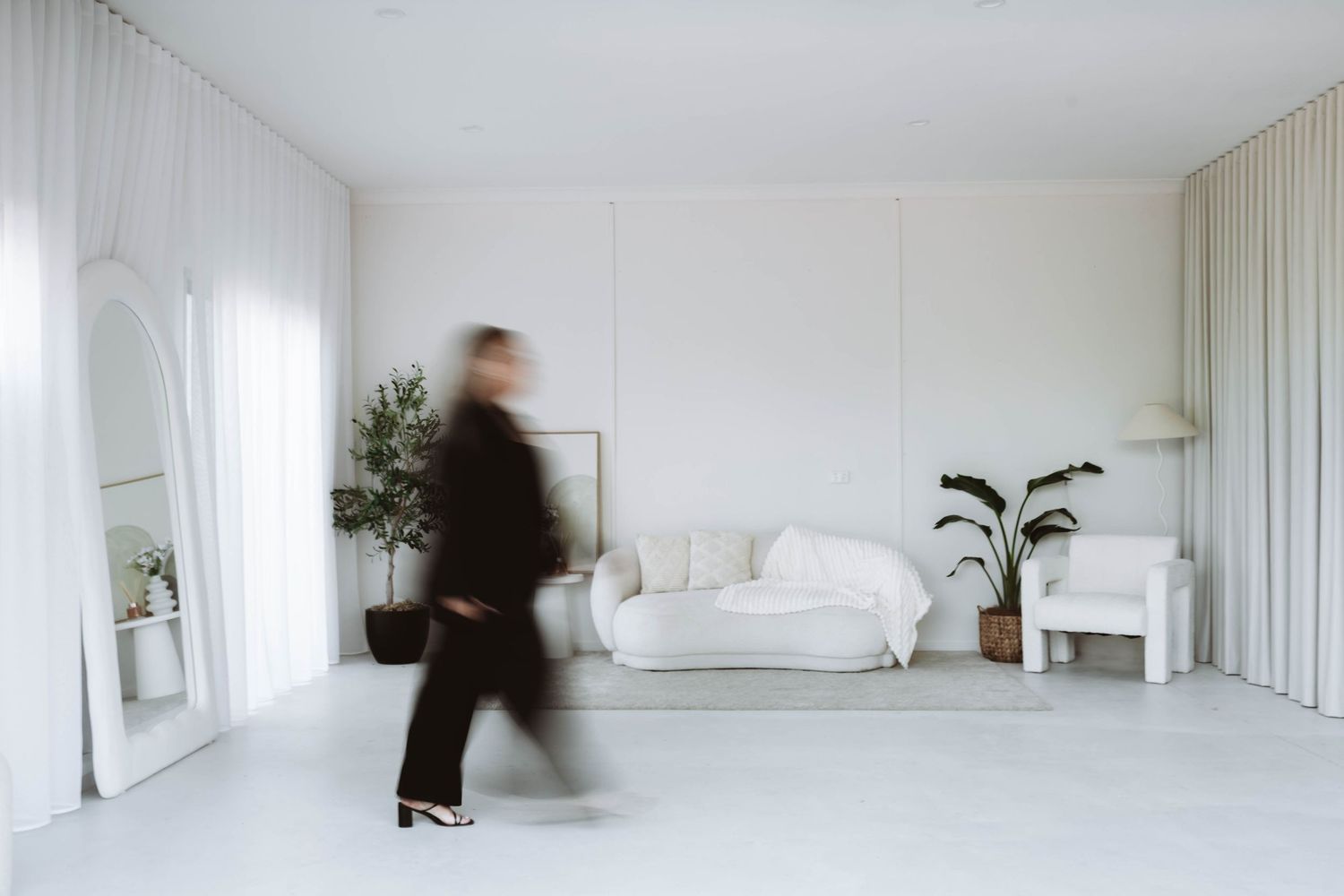 A blurred figure in black walks through a minimalist white living room with a sofa, mirror, and potted plants.