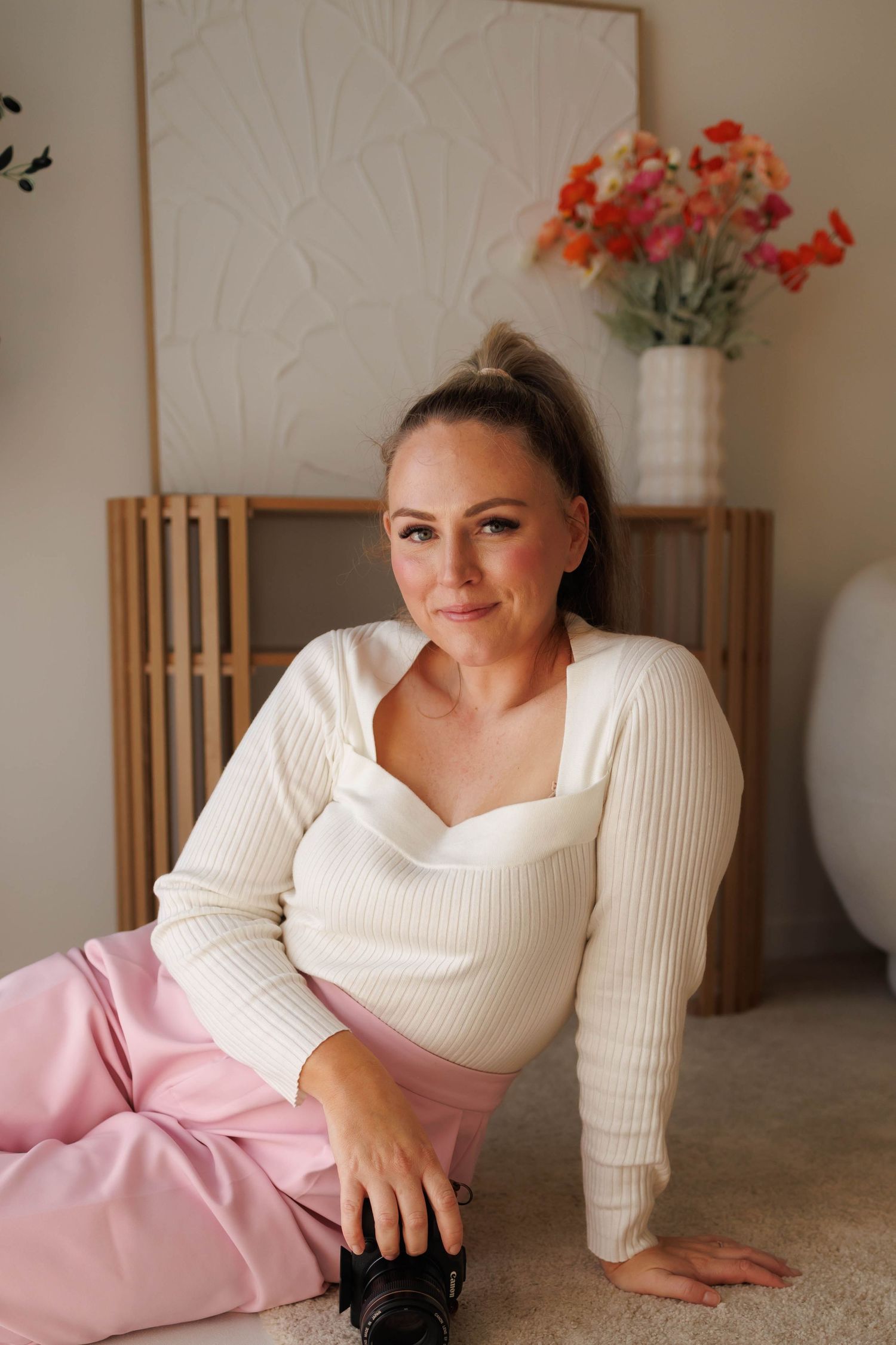 Woman in cream ribbed sweater and pink pants sits on floor holding camera with floral arrangement in background.