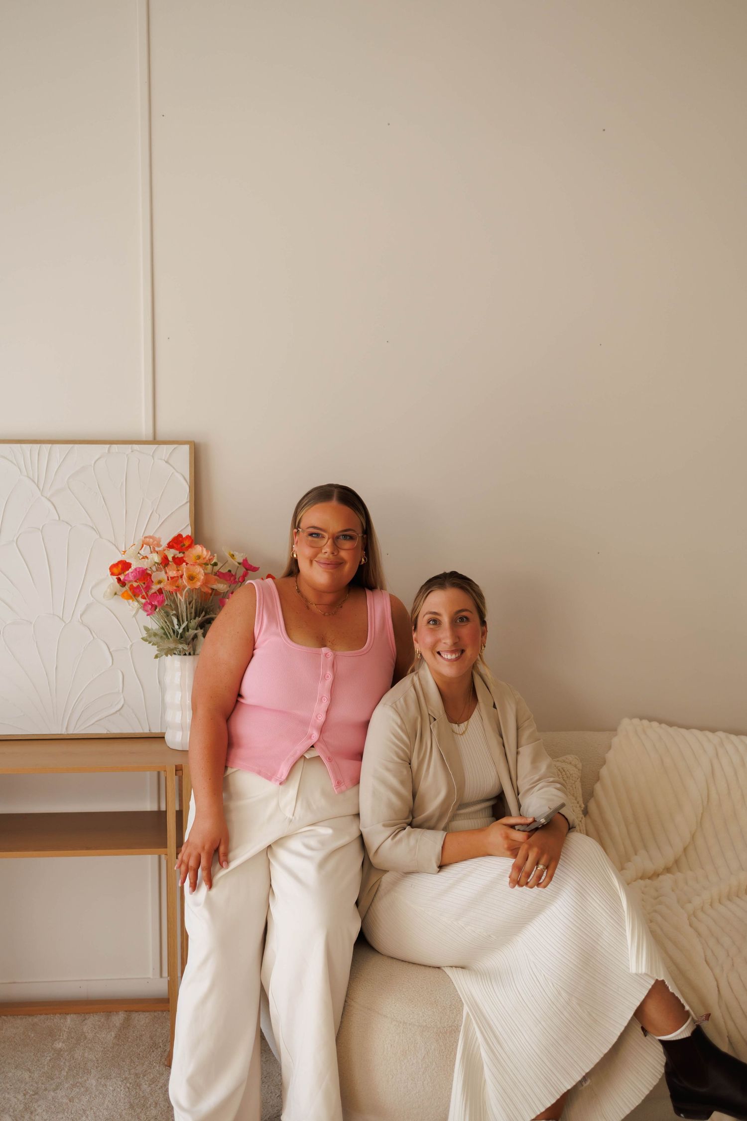 Two women sitting together on a cream sofa, one in a pink top and one in a white blazer, with colorful flowers nearby.
