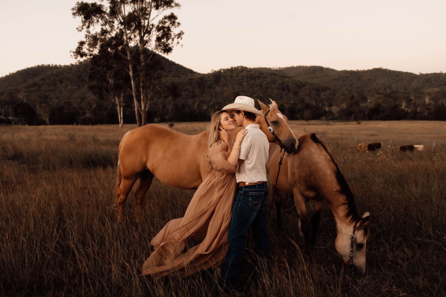 Couple in romantic embrace beside grazing horses in golden field at sunset, woman in flowing dress, man in cowboy hat.