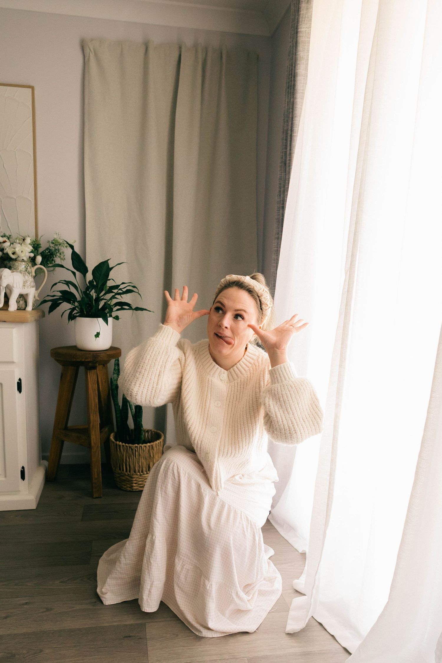 Woman in cream knit sweater and white skirt sits by curtained window, hands raised expressively, indoor plants in background.