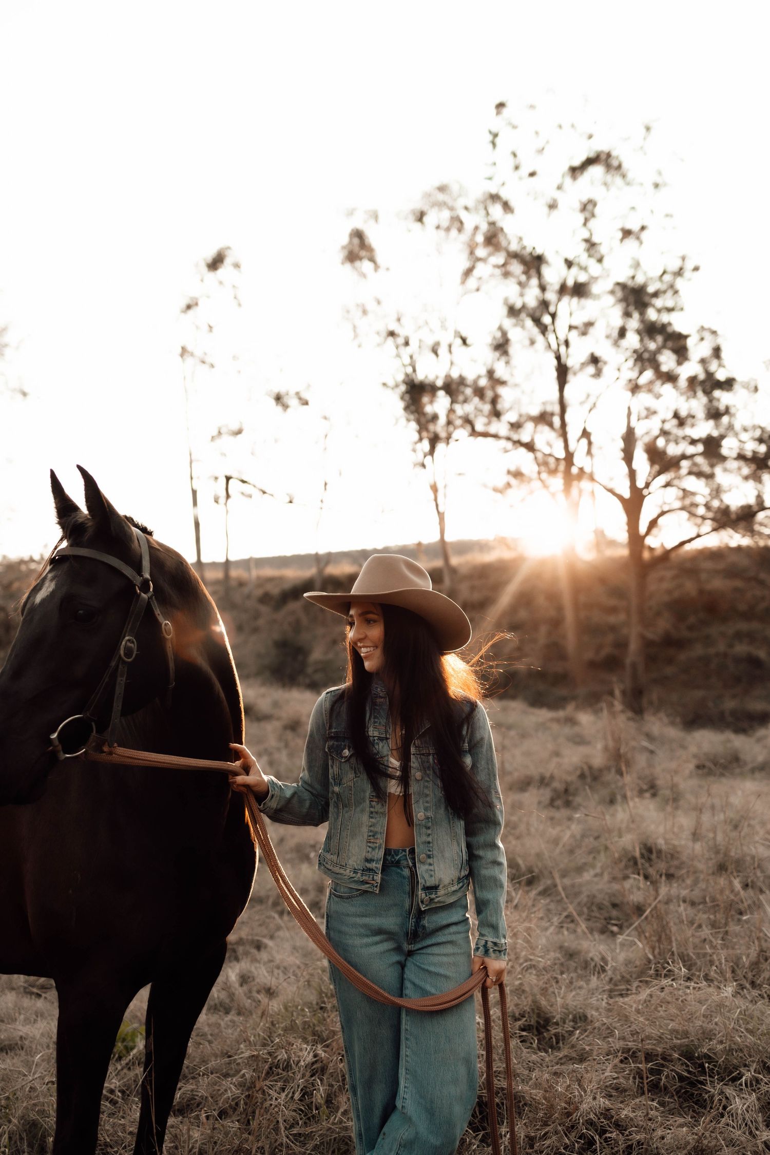 A smiling woman in a cowboy hat and denim jacket leads a dark horse through a golden sunset field.