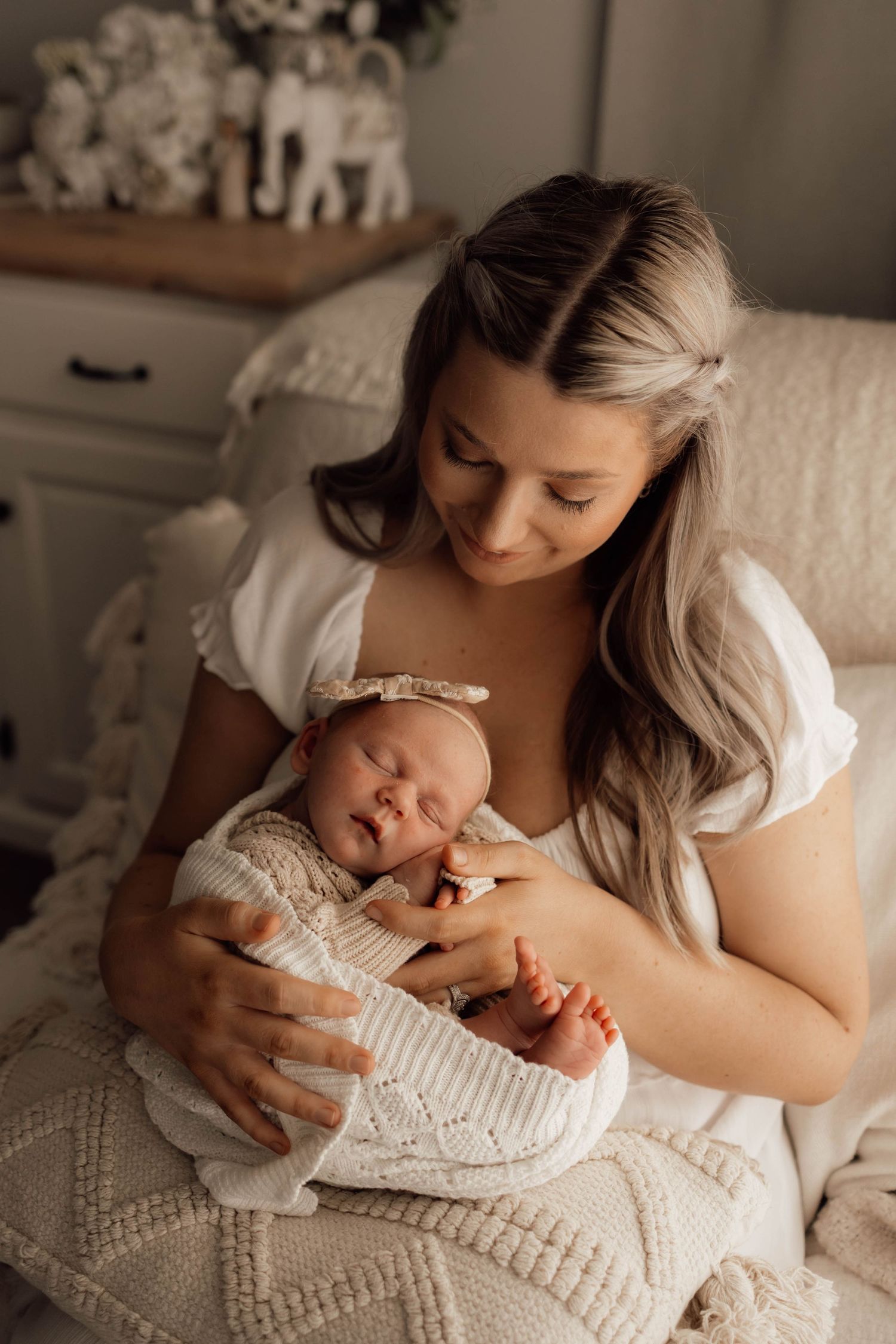 A mother in a white dress tenderly holds her sleeping newborn baby wrapped in a cream knit blanket with a bow headband.