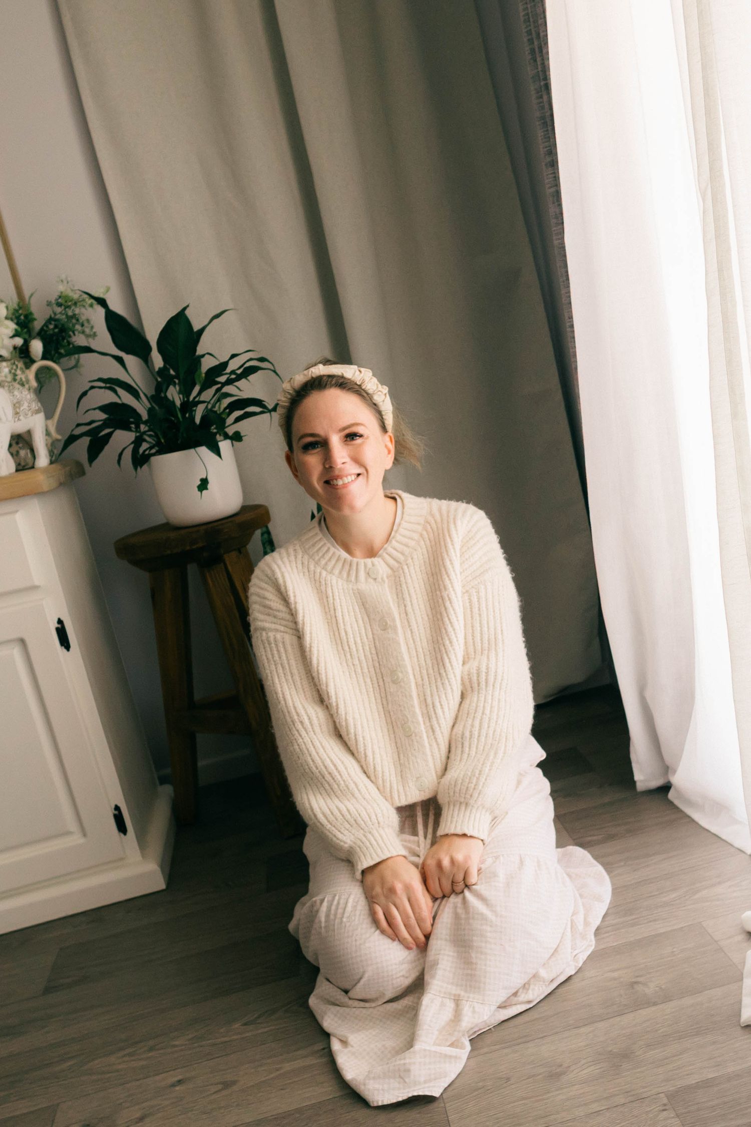 Smiling woman in cream knit sweater sitting on floor near white curtains and green plant.