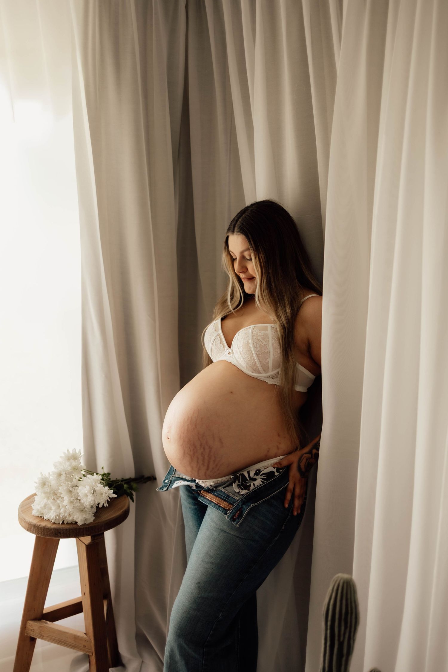 Pregnant woman in white bra and jeans standing by curtains, cradling her baby bump near a wooden stool with white flowers.