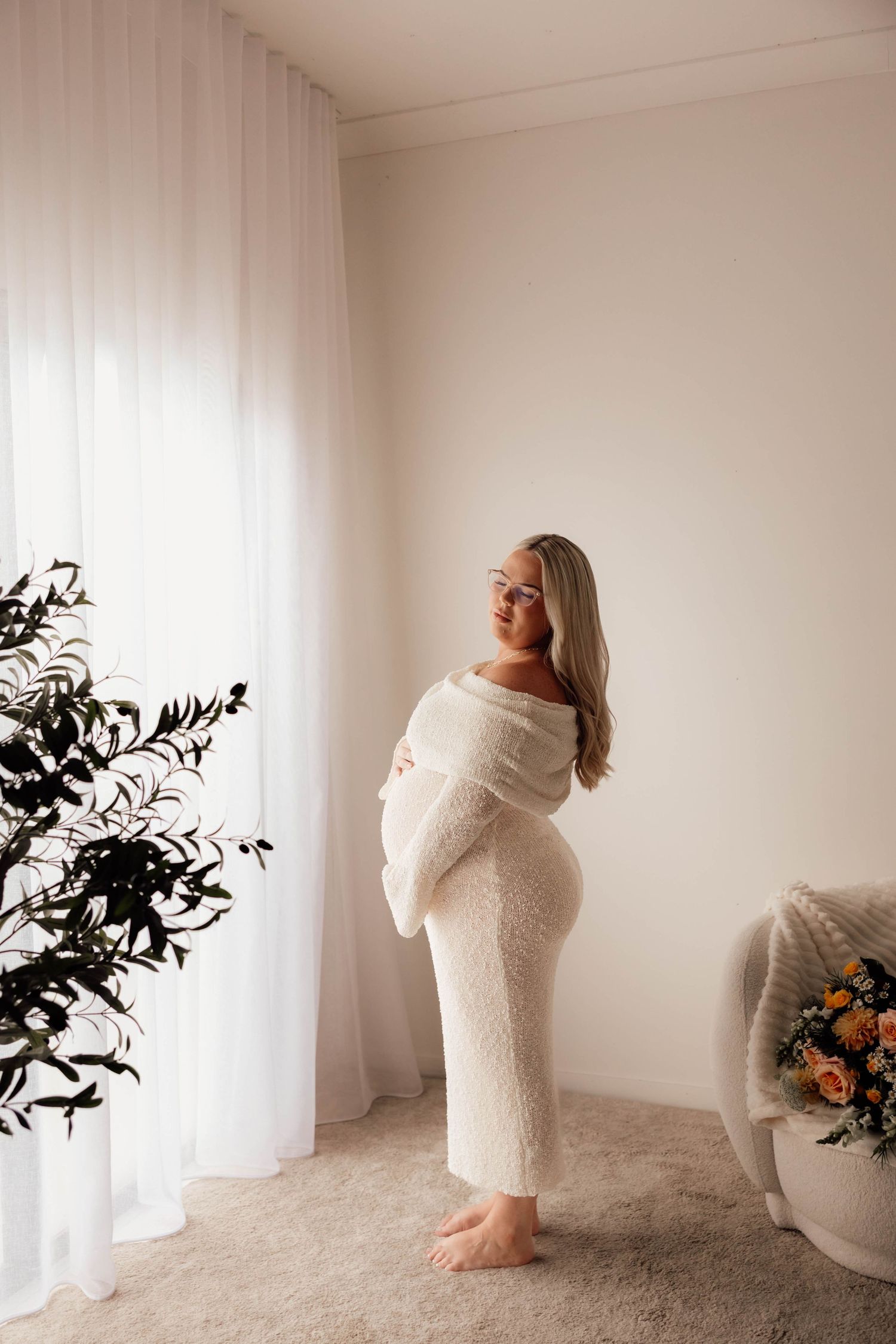 Pregnant woman in white lace dress holding baby bump near sheer curtains in bright minimalist room.