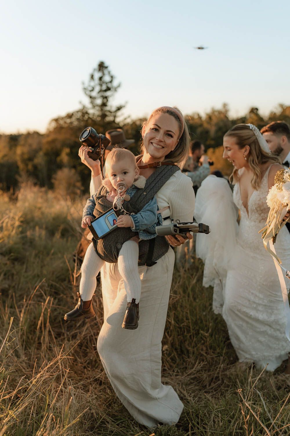 Photographer holding baby in carrier and camera at outdoor wedding golden hour celebration.