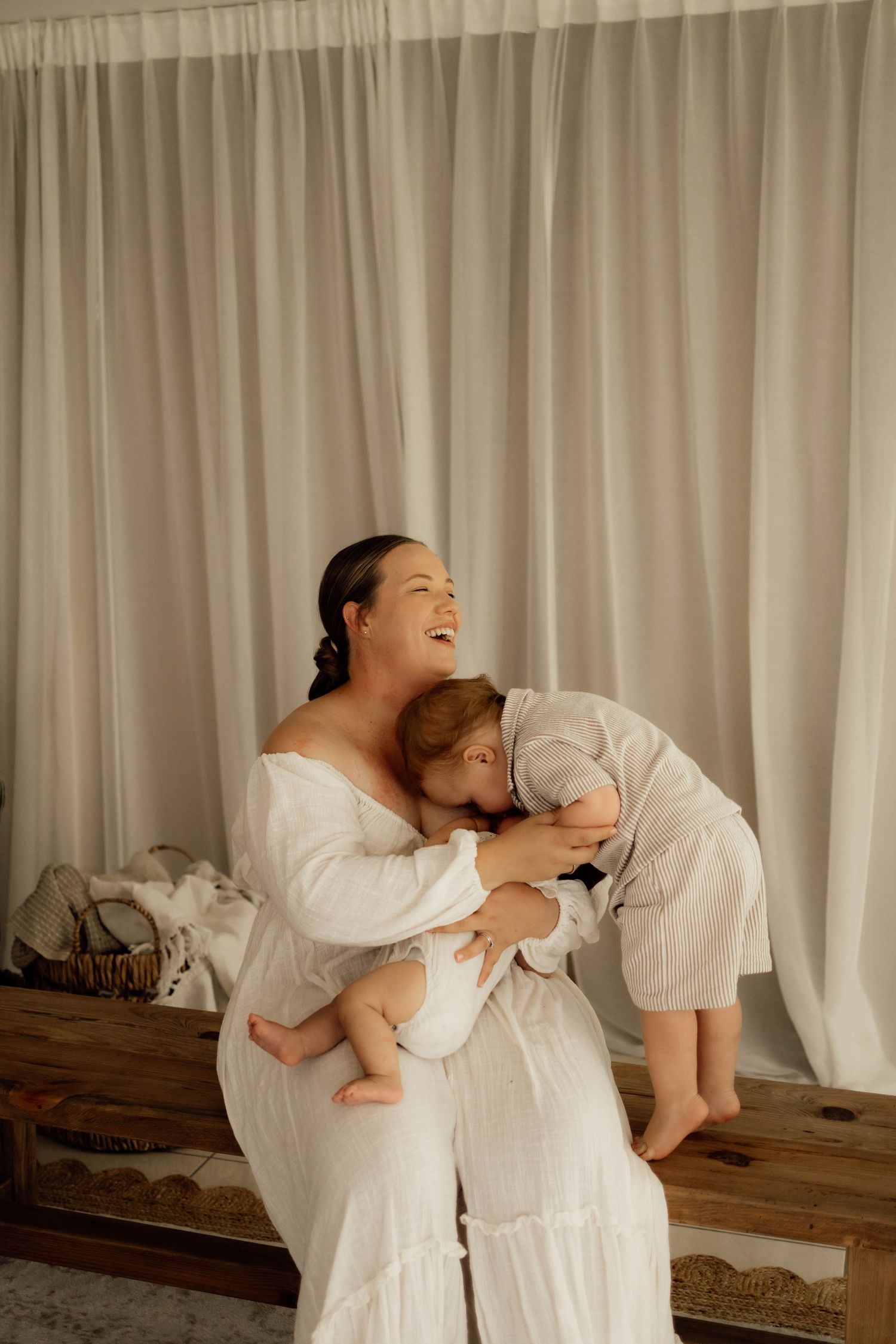 A laughing woman in a white dress holds a baby while a toddler in linen hugs her, posed before sheer white curtains.