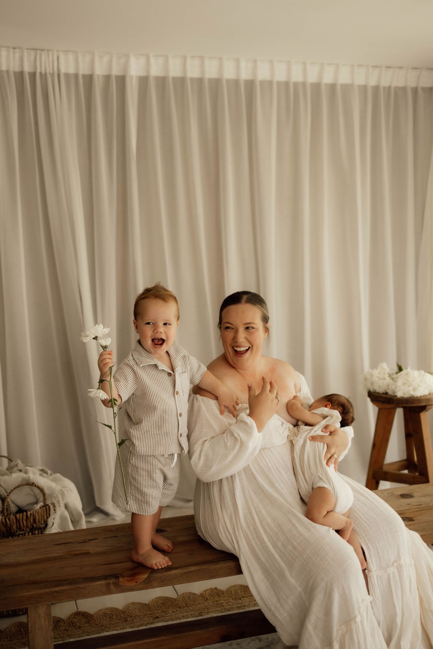 A joyful mother in a white dress sits with her toddler son and newborn baby in a soft, neutral-toned studio setting.