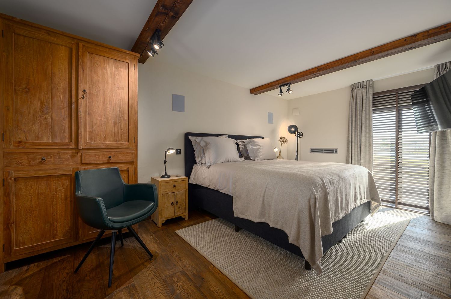 Rustic bedroom with wooden beams, blue accent chair, and built-in wardrobe featuring neutral bedding and natural light, by Parallel Studios