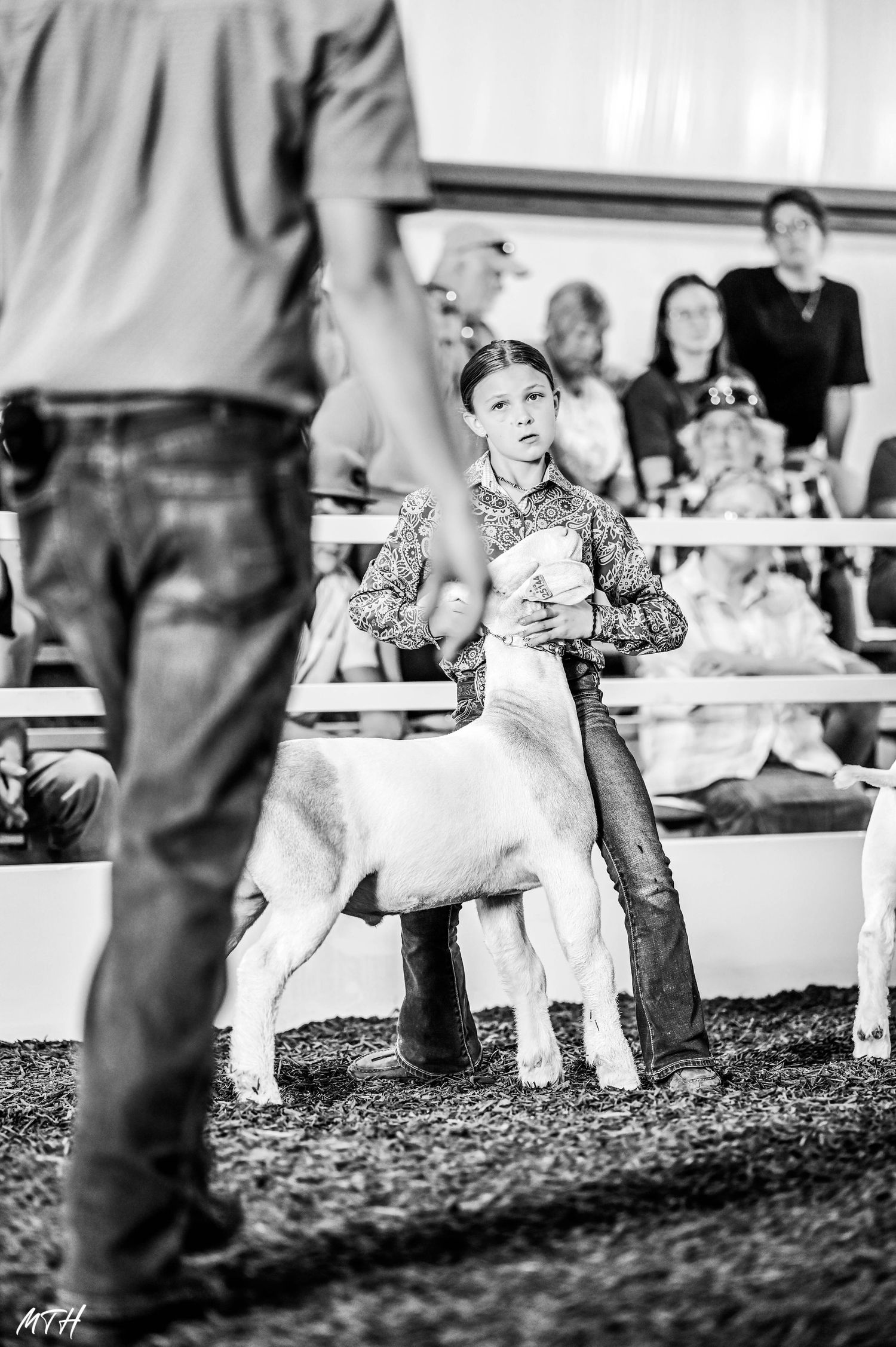 Black and white photo of a youth showing a white goat at a livestock competition in a dirt arena.